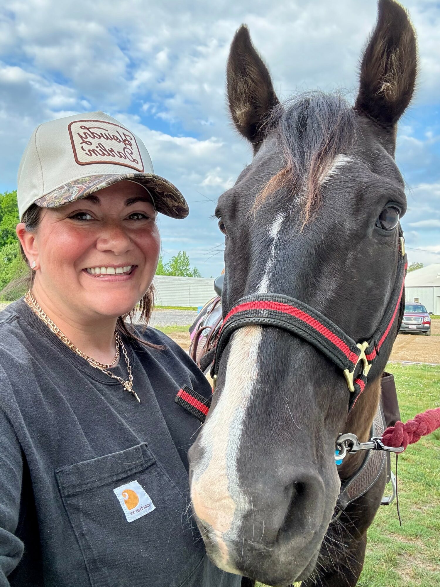 Person smiling next to a black horse outdoors with a blue sky and clouds, wearing a cap and dark shirt.