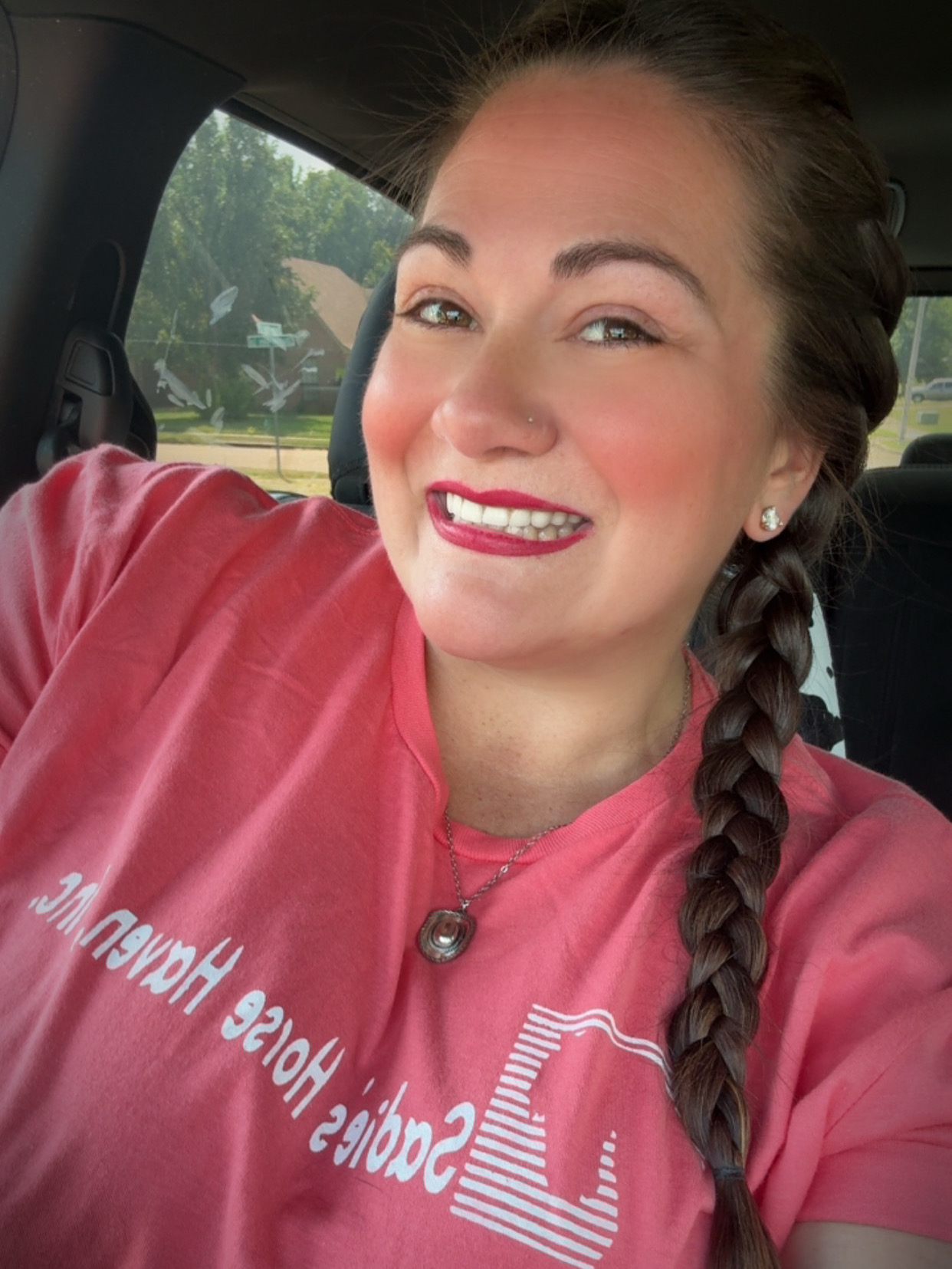 Smiling woman with braided hair in a car, wearing a pink shirt and a necklace, with trees visible outside.