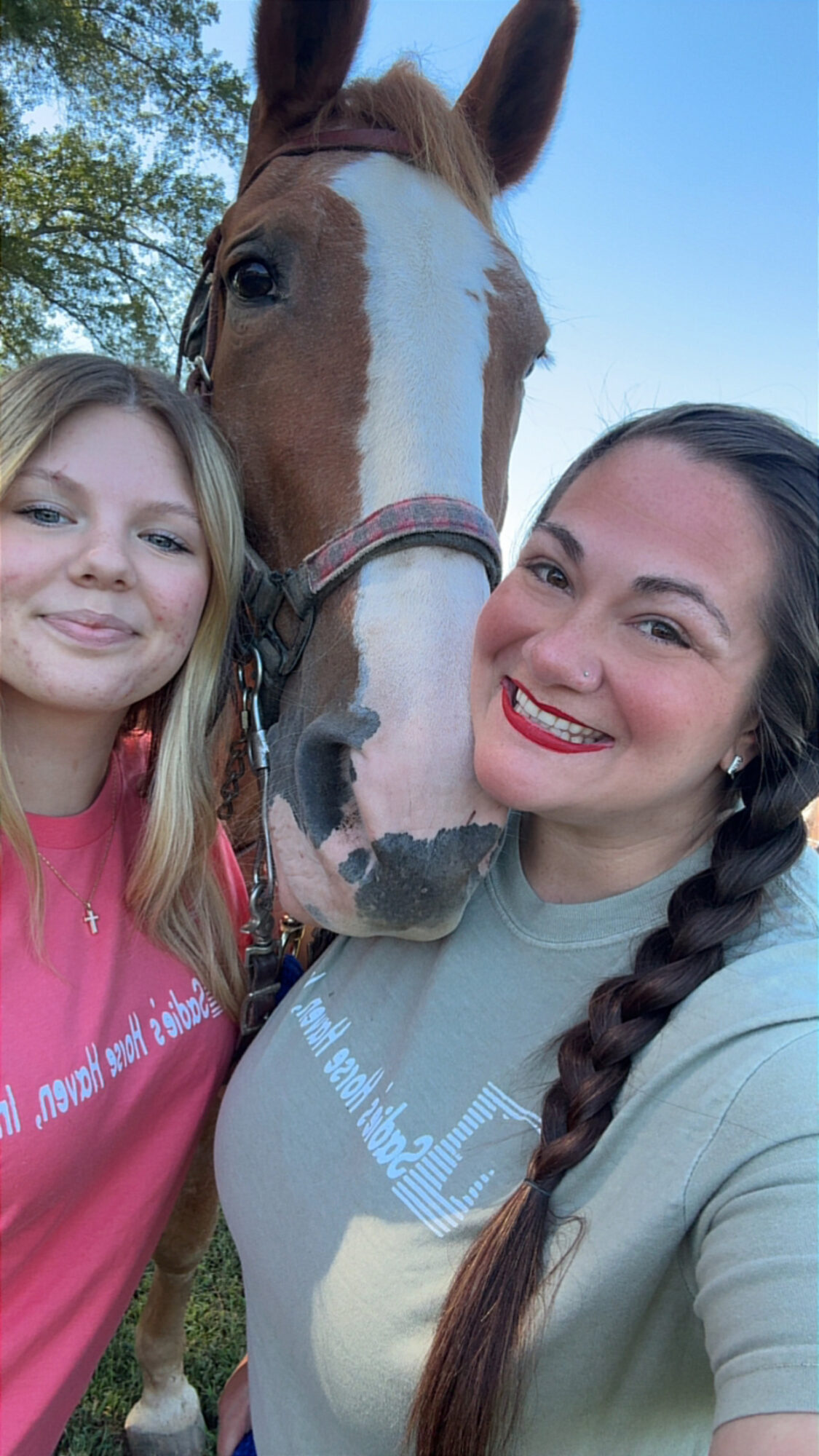 Two women and a horse outdoors, smiling at the camera, with trees and blue sky in the background.