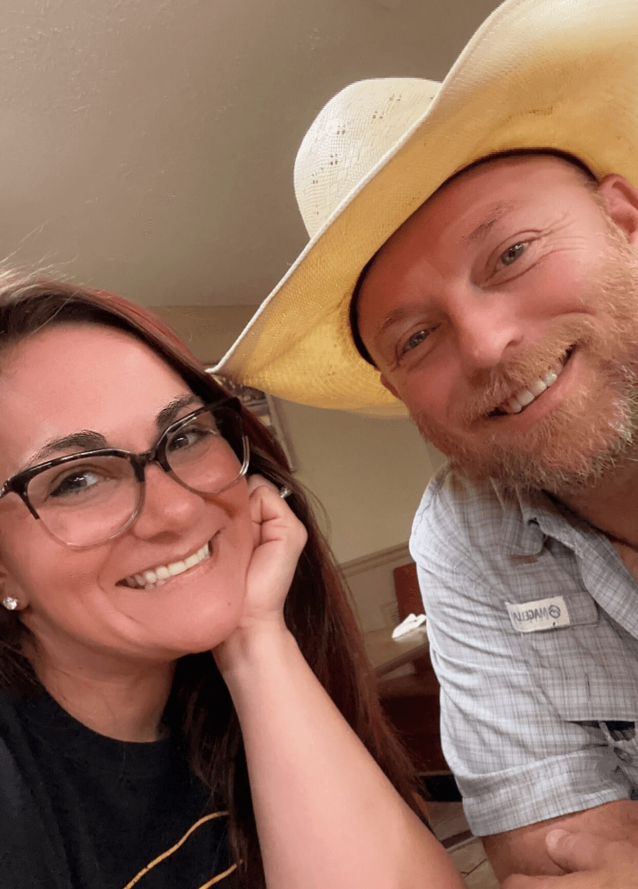 Two smiling people, one wearing glasses and the other wearing a cowboy hat, close together indoors.
