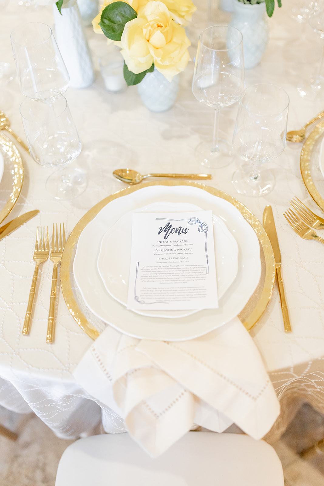 Elegant table setting with a white plate, gold cutlery, a menu, and a white napkin, with floral centerpieces in the background.