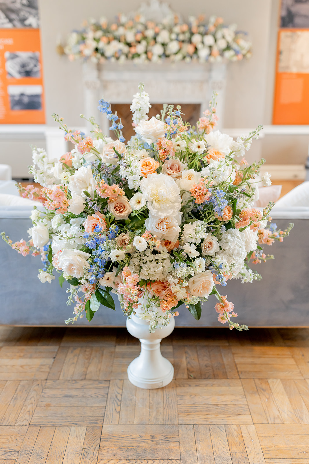 Large floral arrangement in a white vase with roses, peonies, and other flowers in pastel colors, on a wooden floor.