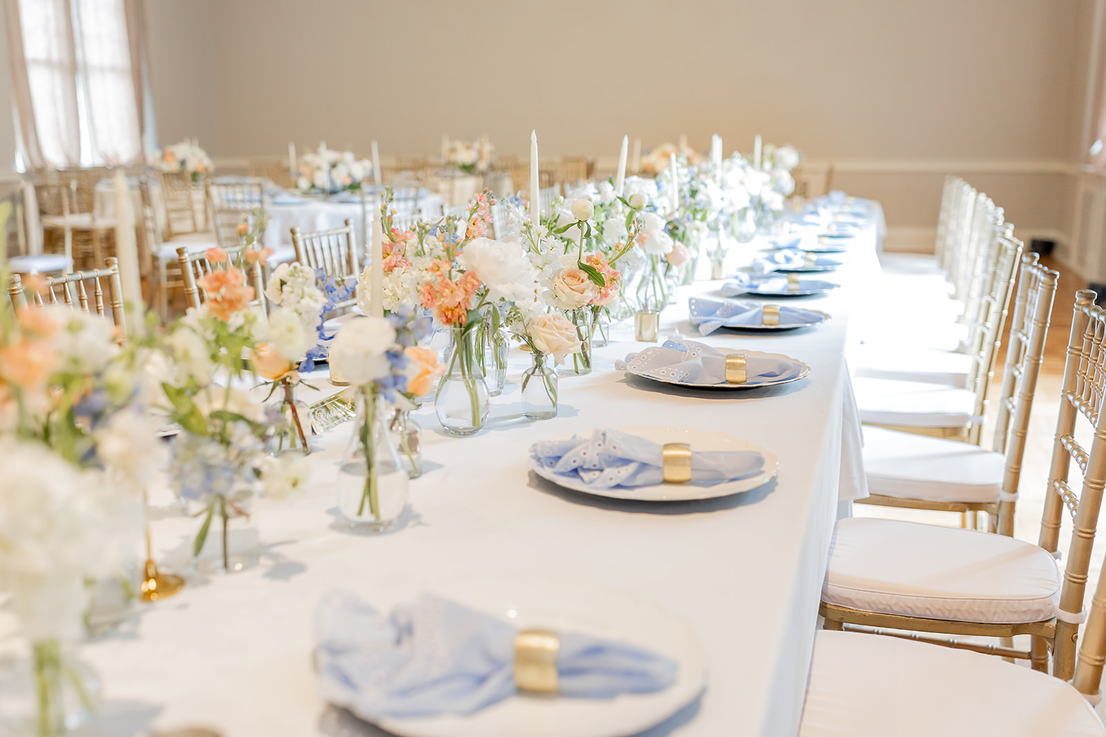 Long banquet table set with plates, napkins, and floral centerpieces in a bright room.