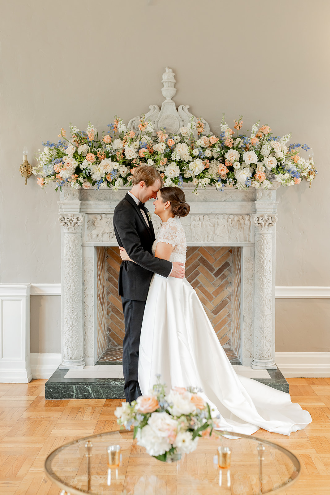 A bride and groom stand close in front of a decorated fireplace, holding hands and looking at each other, with flowers on the floor.