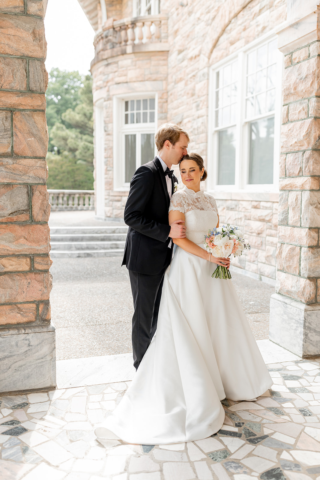 Bride and groom standing outside a stone building, the groom kissing the bride's forehead, she holds a bouquet, sunlight filters through windows.