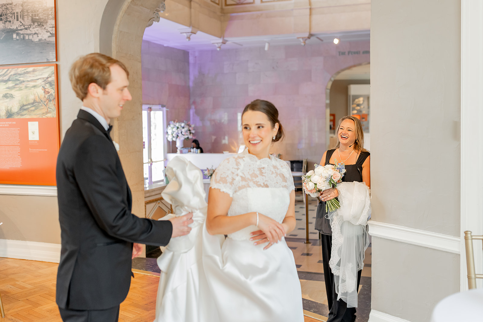 Bride and groom smiling at each other during wedding reception, woman holding bouquet, woman in background smiling, indoor setting