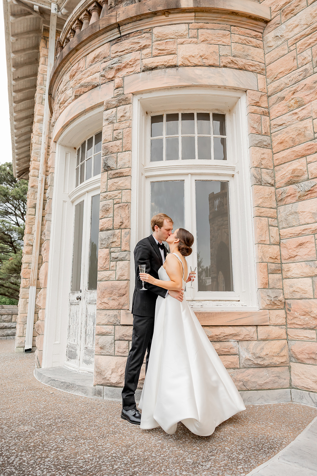 A couple in wedding attire standing close outside a stone building with large windows, embracing and looking at each other.