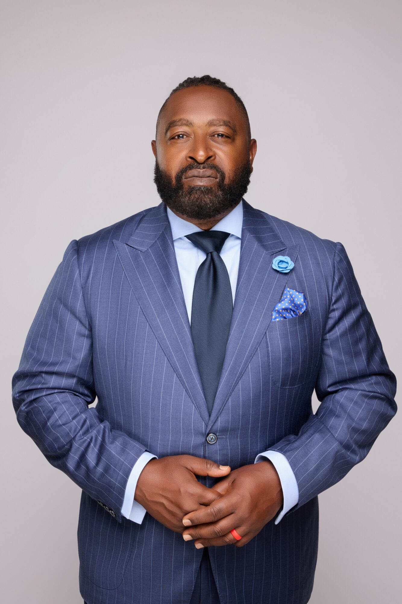 Man in a blue pinstripe suit with a tie and pocket square, standing against a plain background.