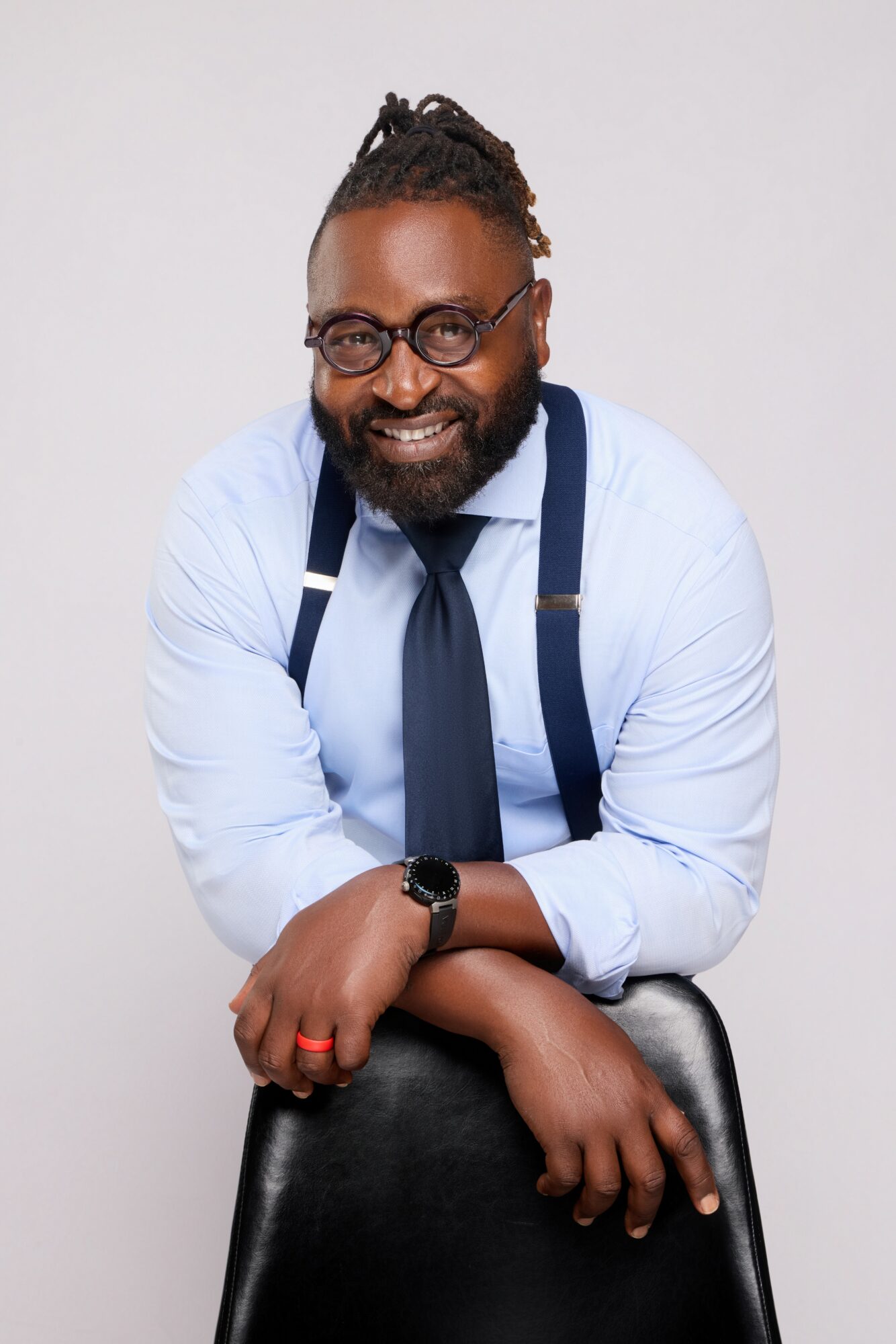 Smiling man with glasses, beard, and dreadlocks, wearing a light blue shirt, dark tie, and suspenders, sitting on a chair.
