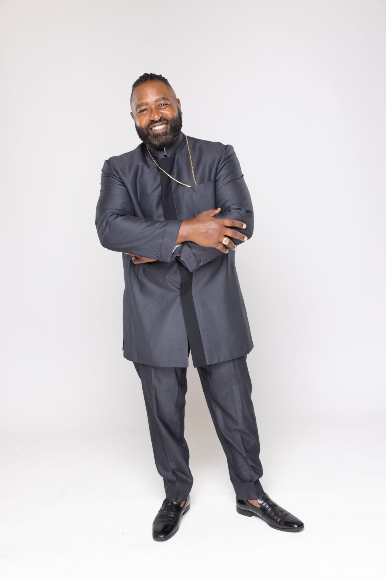 Man with a beard smiling, wearing a dark suit, standing with arms crossed against a plain white background.