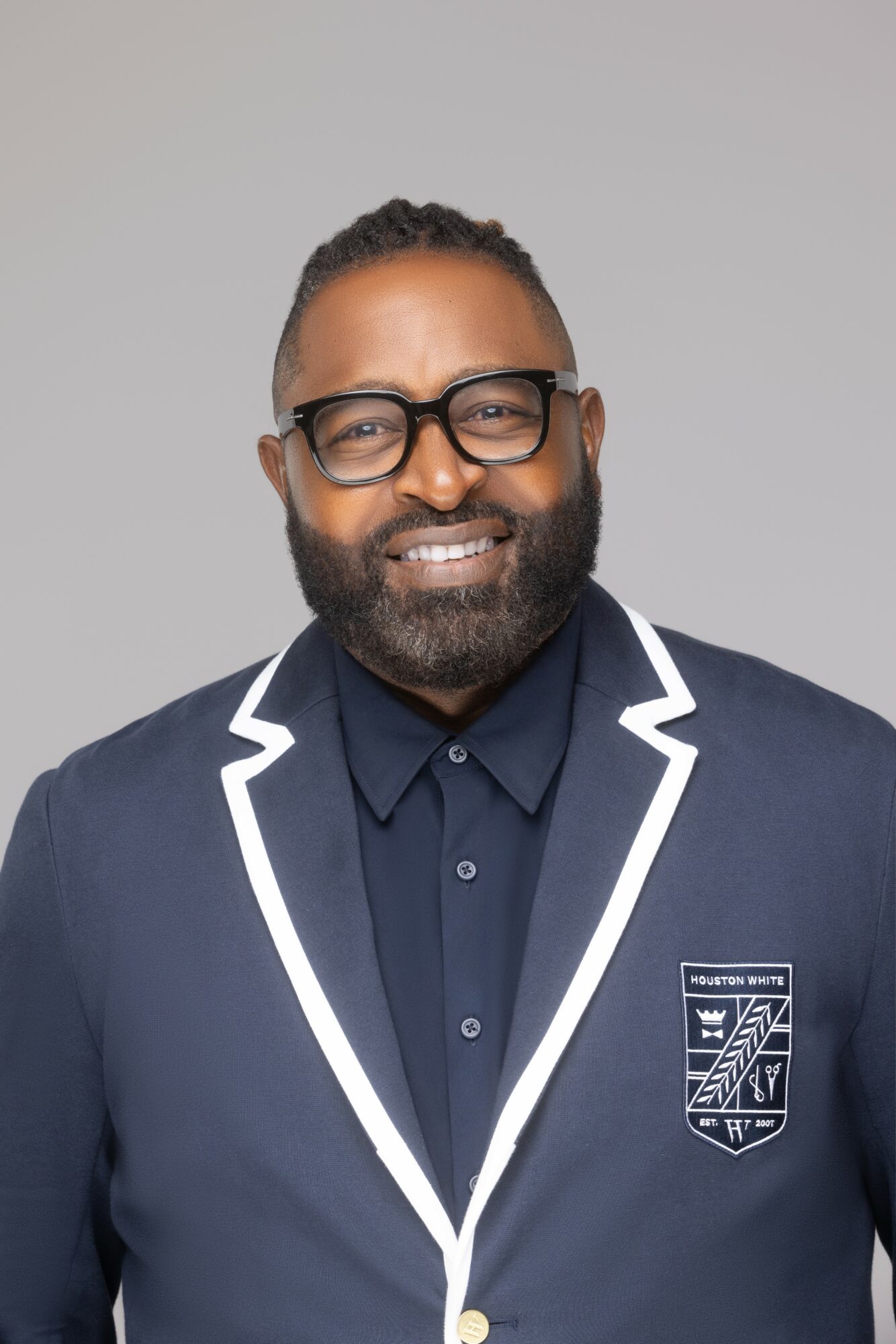 Man with glasses, beard, smiling, wearing a dark blazer with white trim and a crest, against a plain background.