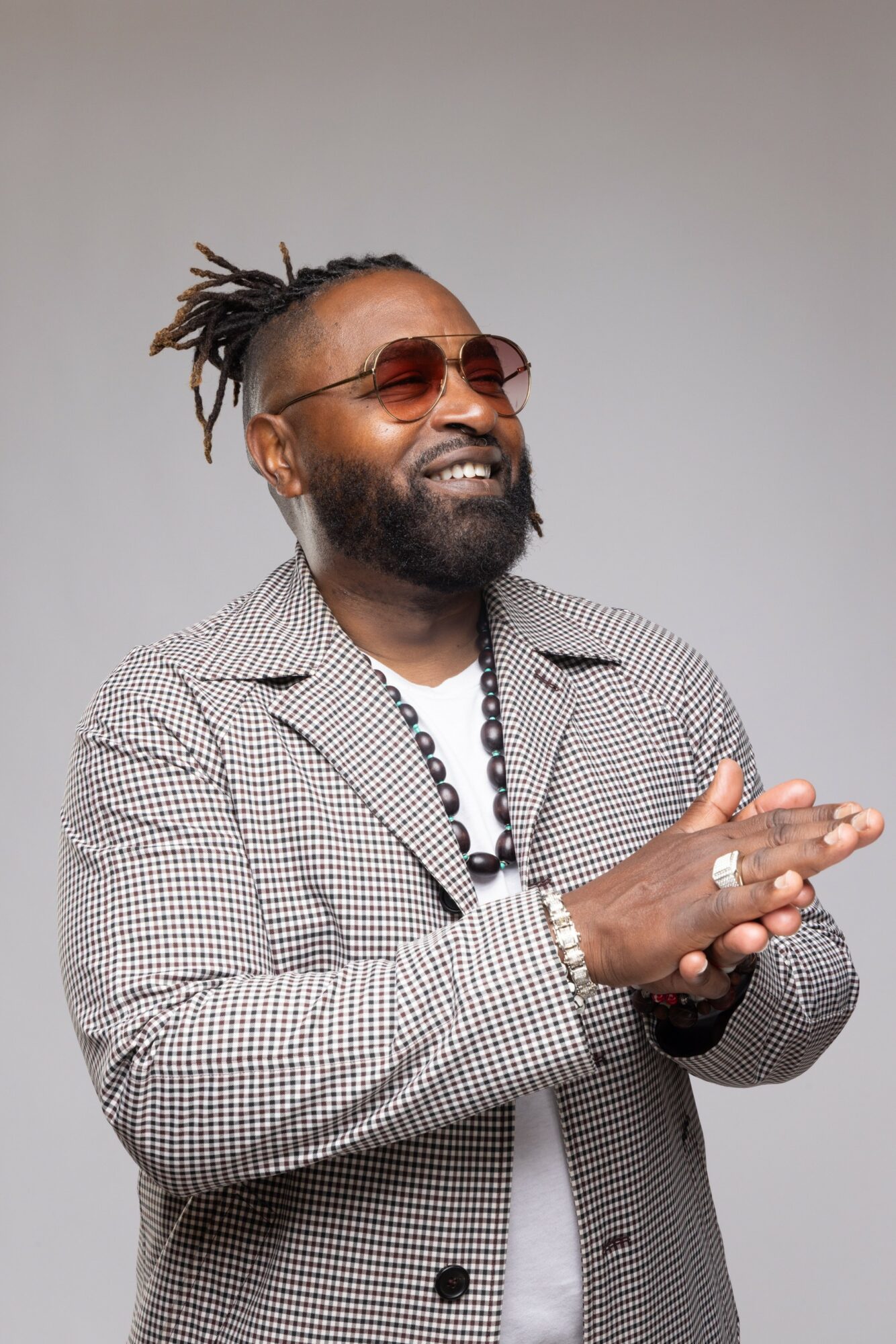 Man with dreadlocks, beard, sunglasses, smiling, wearing a checkered blazer and necklace, clapping hands, against a plain background.