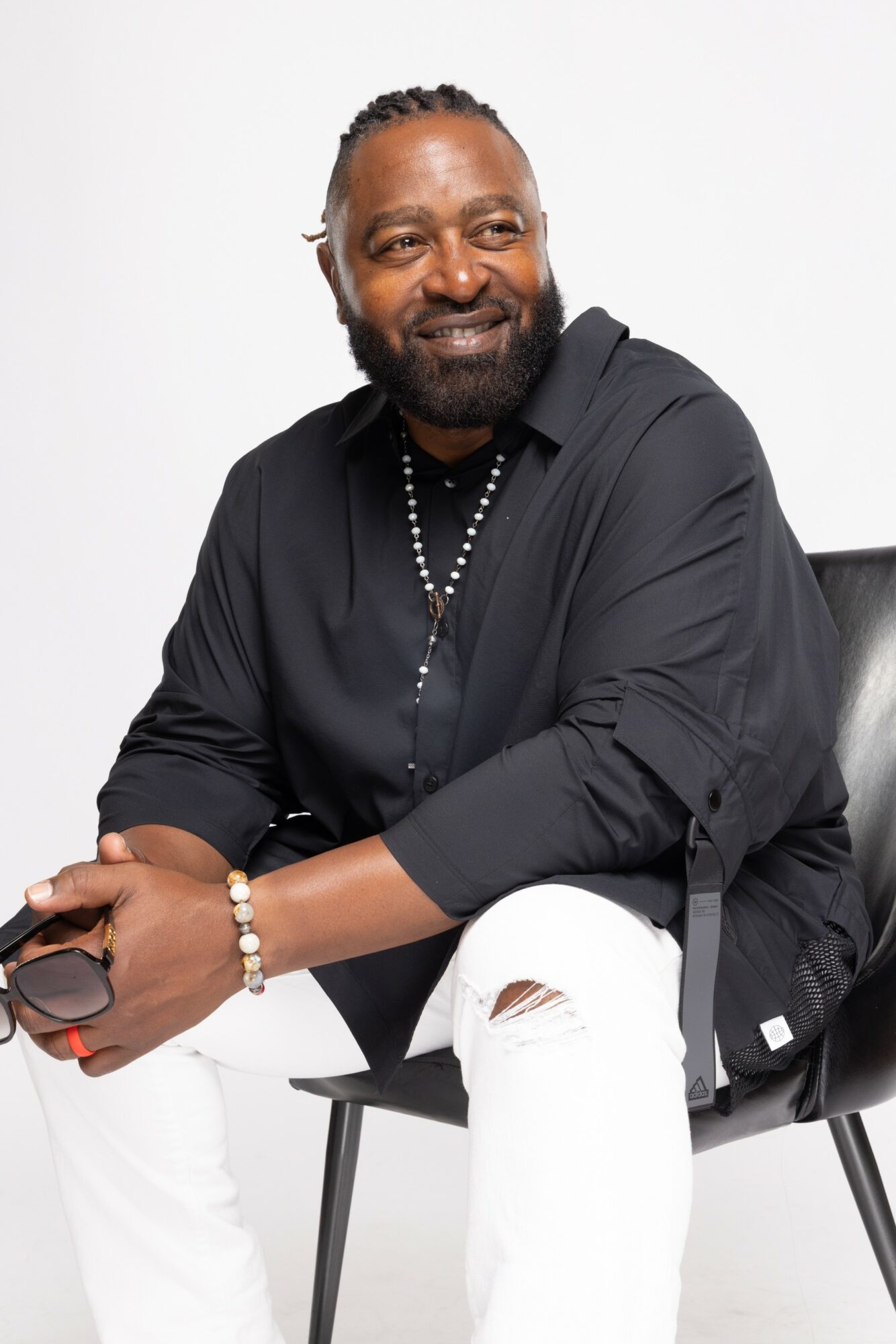 Man with beard and braided hair, smiling, wearing black shirt and white pants, sitting on a chair against a white background.