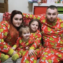 Four people sitting on a couch wearing matching red pajamas with holiday patterns, smiling at the camera.