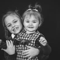 Two young girls smiling, one hugging the other, both with light-colored hair, in black and white.