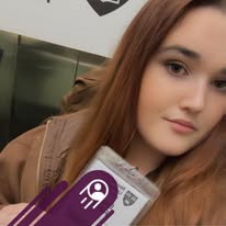 Young woman with long brown hair, wearing a brown jacket, holding a phone, in an indoor setting.