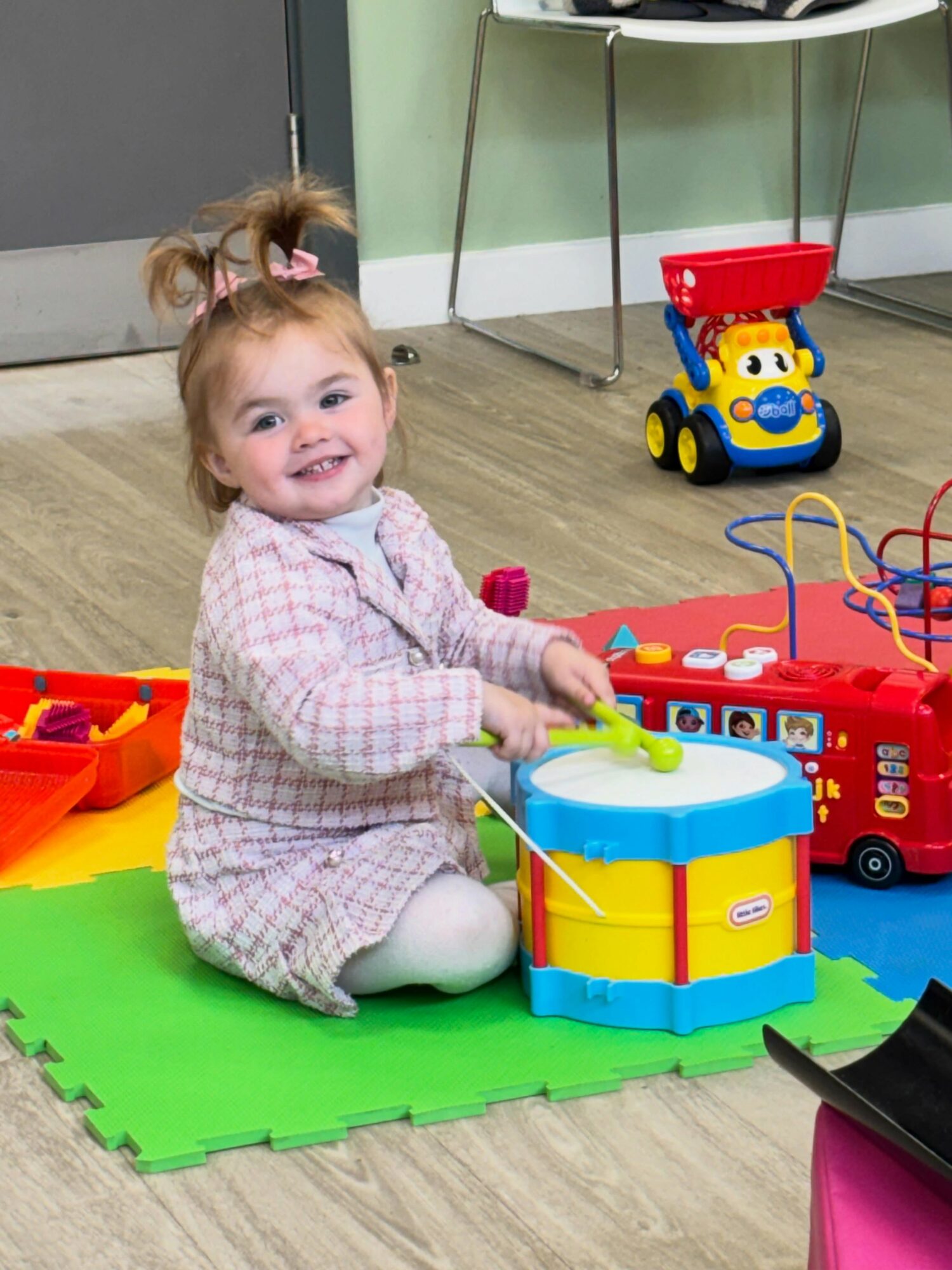 Smiling young girl playing with a drum toy on a green mat in a playroom, surrounded by toys.