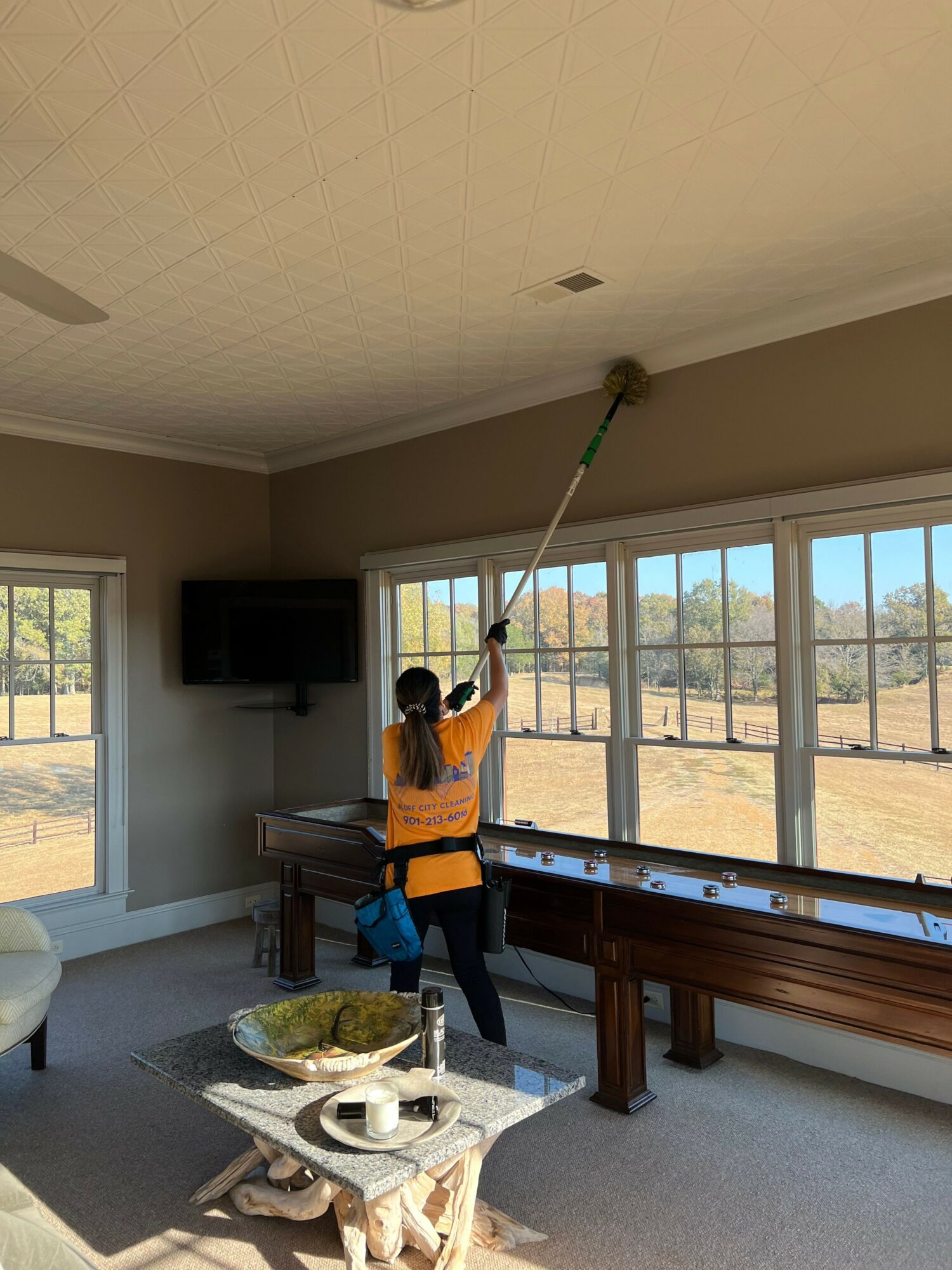 Person cleaning window with a long tool in a room with large windows, a table, and a TV, sunlight streaming in.