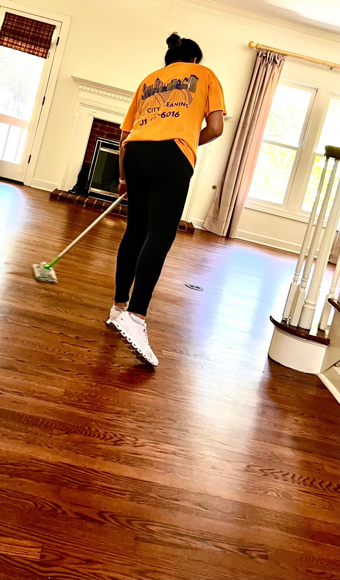 Person mopping wooden floor in a bright room with large windows and curtains, wearing an orange shirt and black pants.