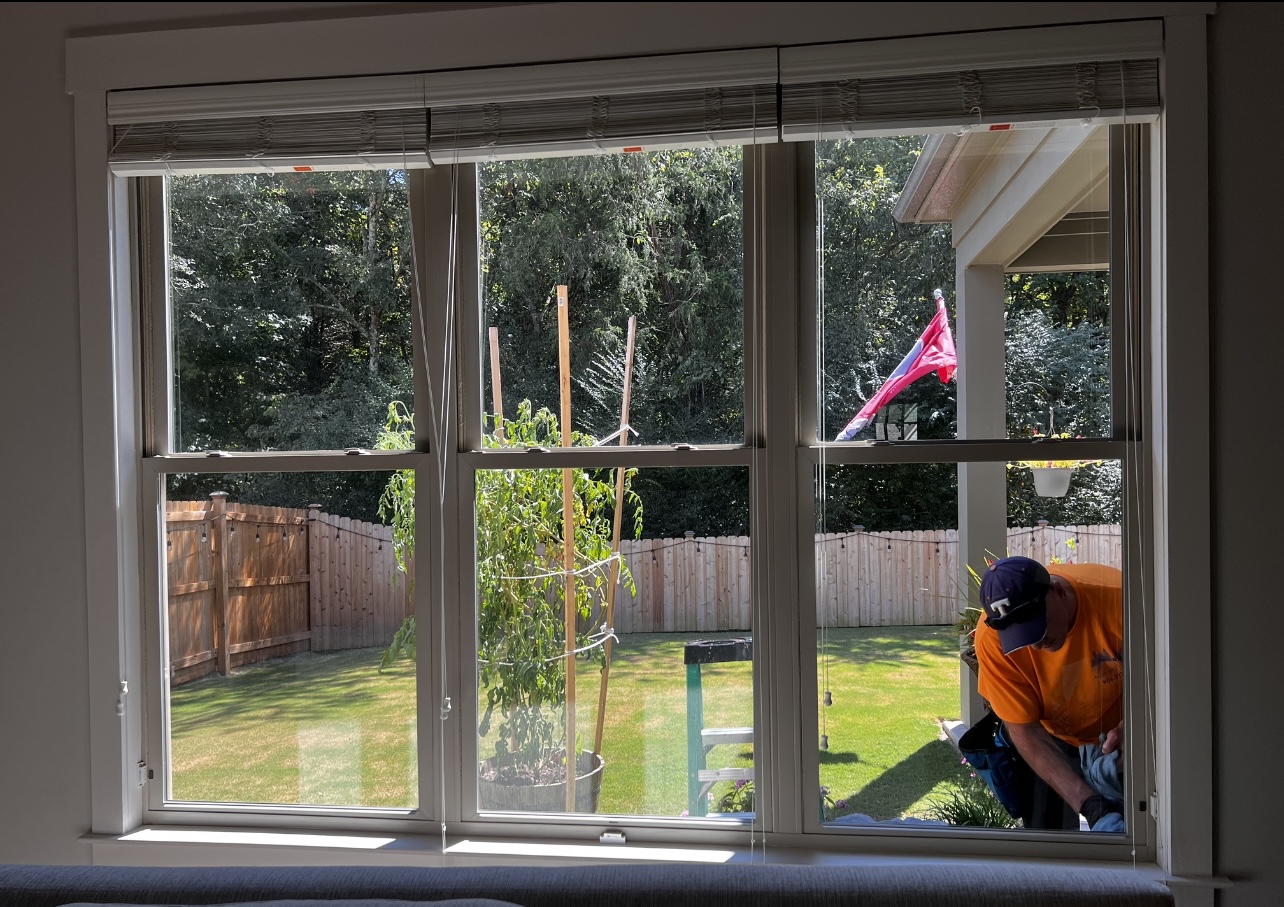 Person in orange shirt working outside near a window, with a backyard, trees, and a pink flag visible.