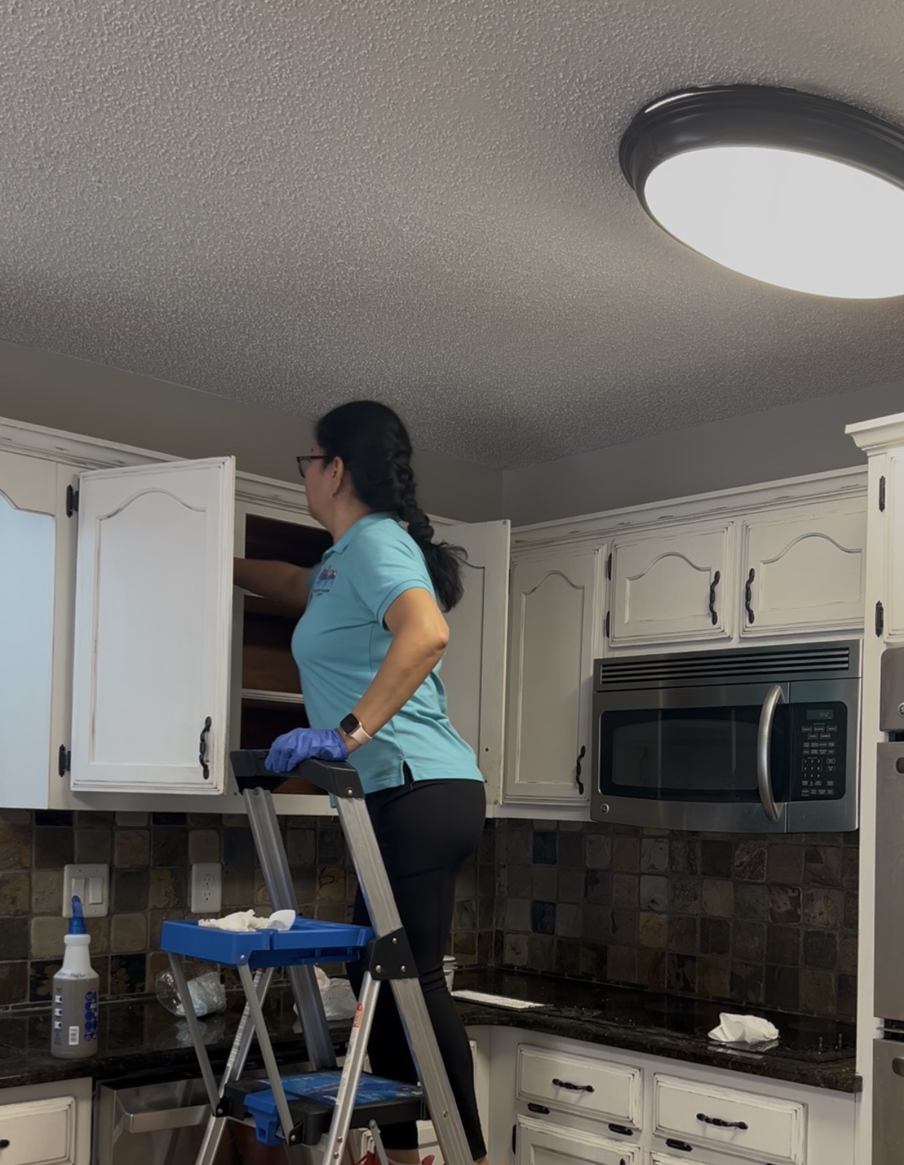 Person standing on a step stool opening kitchen cabinet, with microwave and tiled backsplash visible.