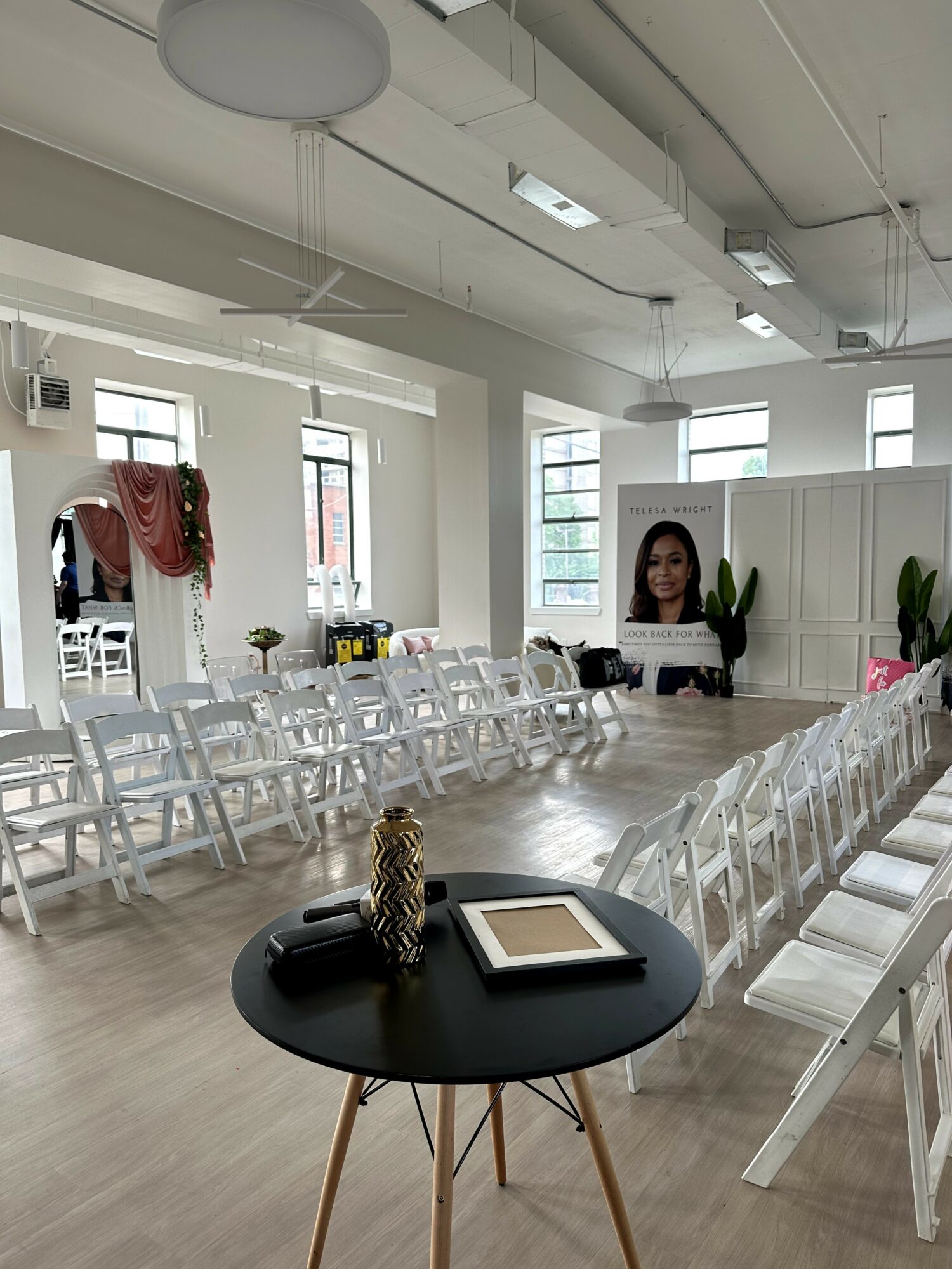 Large room with rows of white chairs, a black table with decorative items, and a woman on a screen, bright windows.