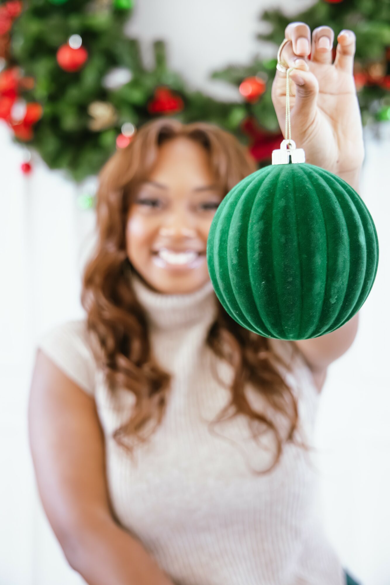 Person holding a green Christmas ornament in front of a decorated tree, smiling, with blurred background.
