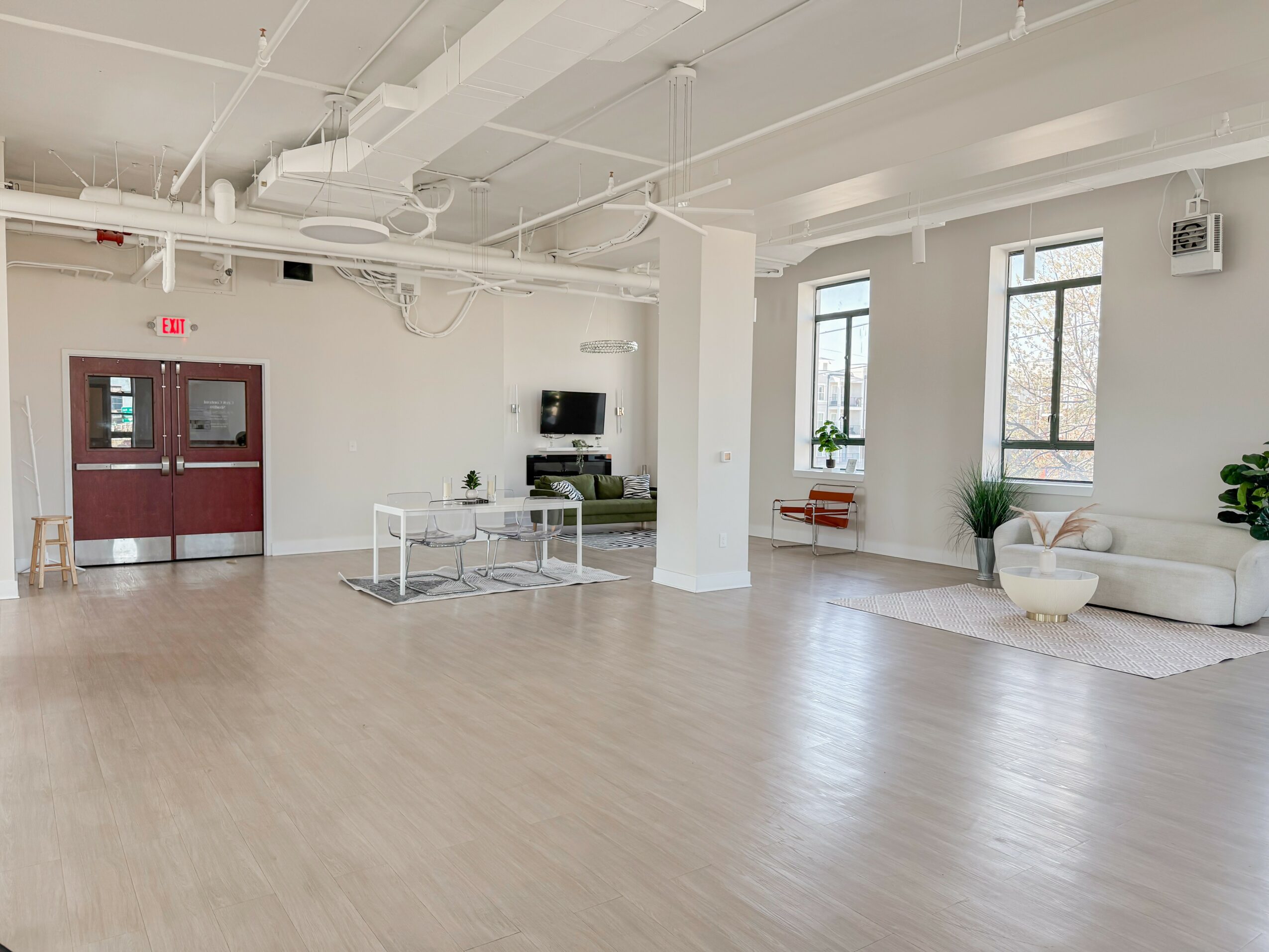 Spacious room with large windows, white walls, light wood flooring, and minimal furniture including a sofa, plants, and a small table.