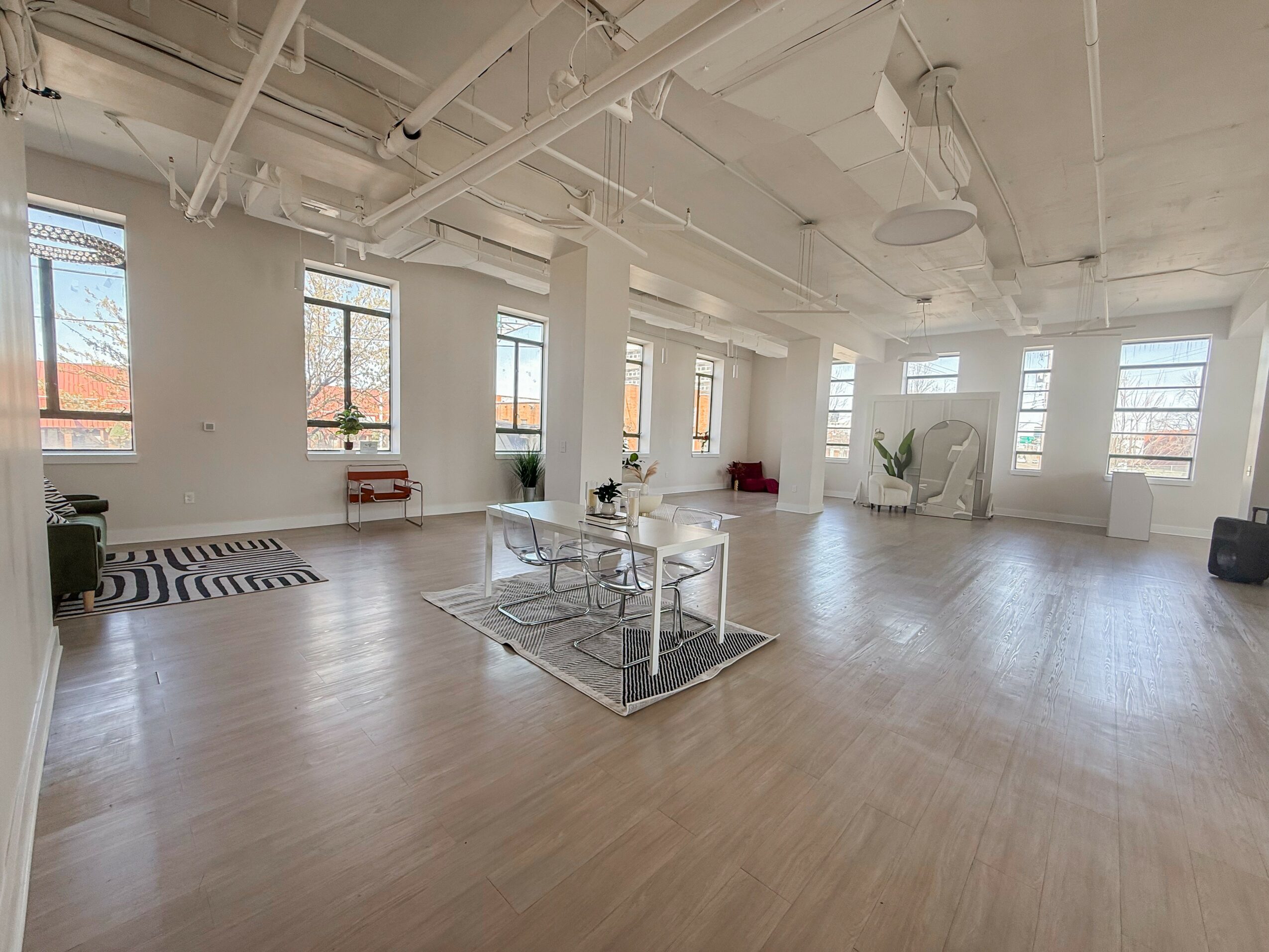 Spacious room with large windows, white walls, light wood flooring, and minimal furniture including a glass table and plants.