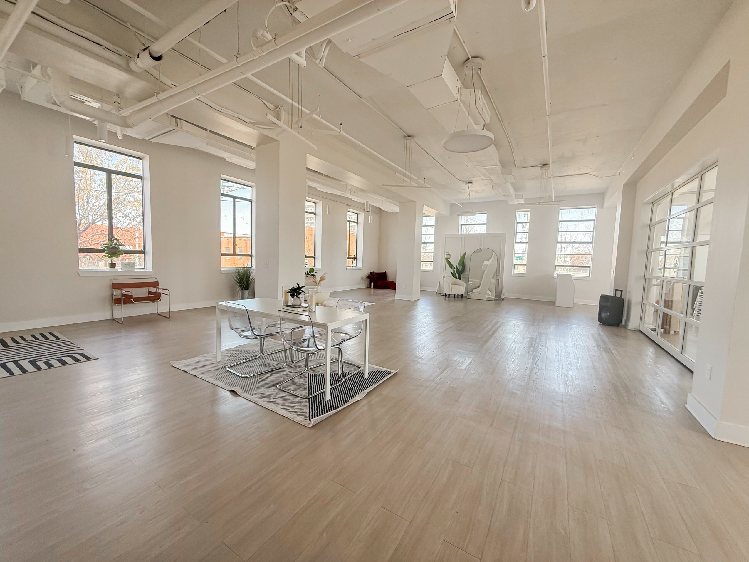Spacious room with large windows, white walls, light wood flooring, and minimal furniture including a table, chairs, and plants.