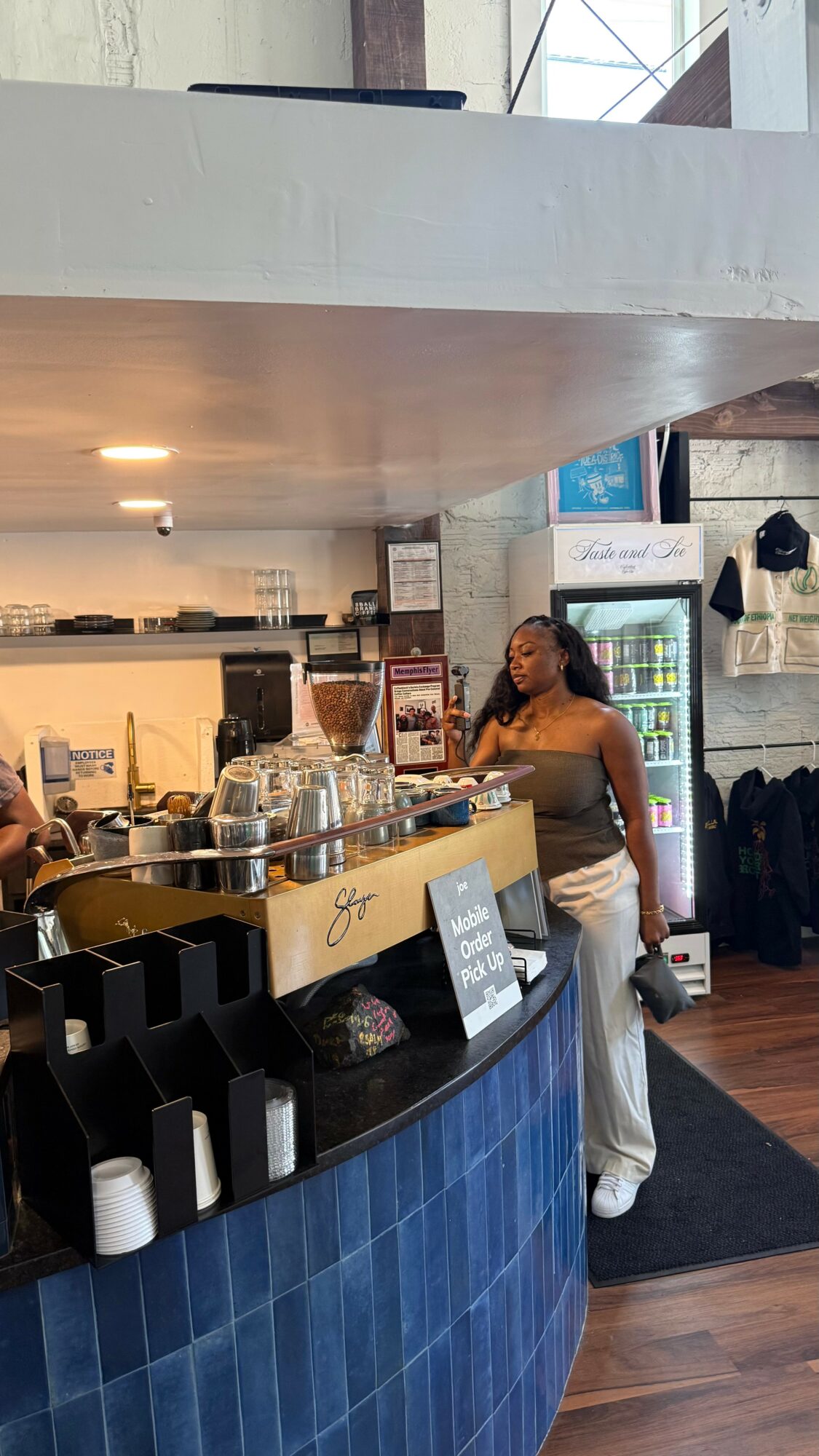 A woman stands at a counter in a cafe, with a refrigerator and shelves in the background.