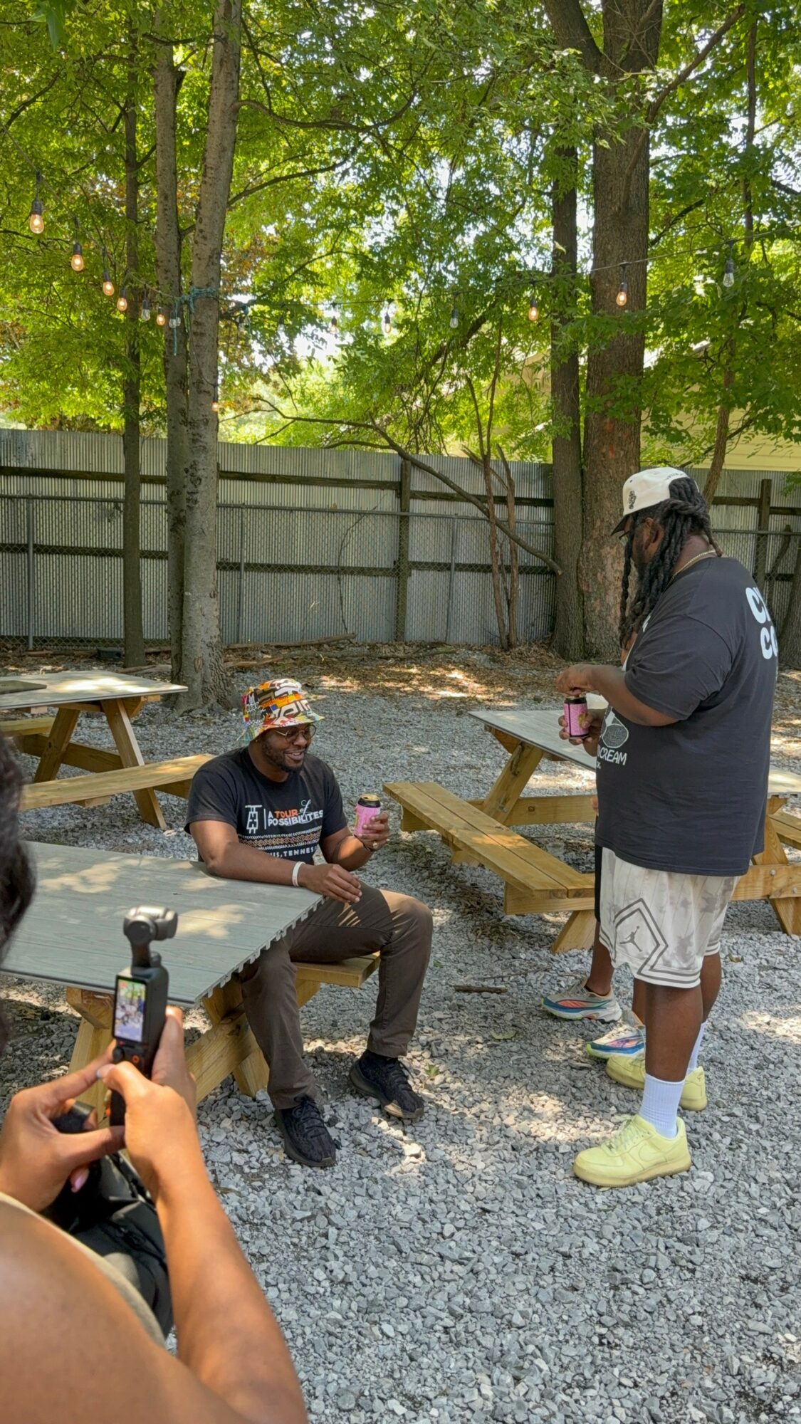 Two people outdoors near trees and a wooden fence, one sitting and the other standing, with picnic tables nearby.