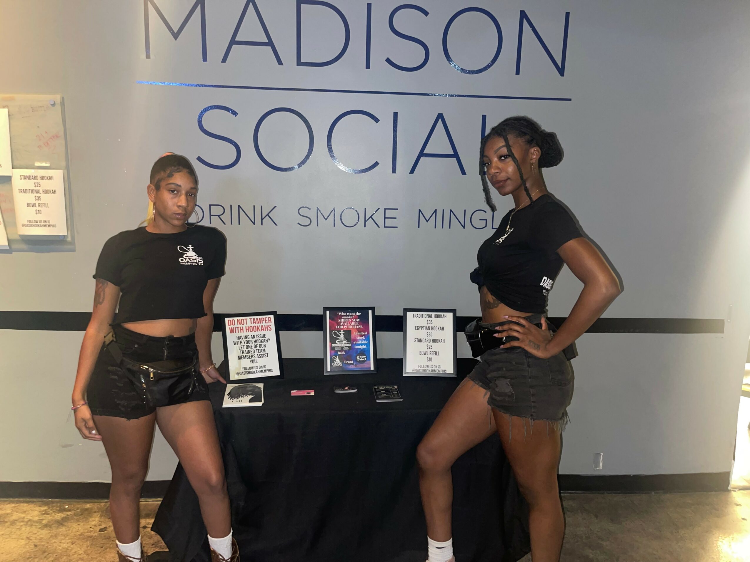 Two women stand on either side of a table with digital screens and signs, in front of a wall with text.