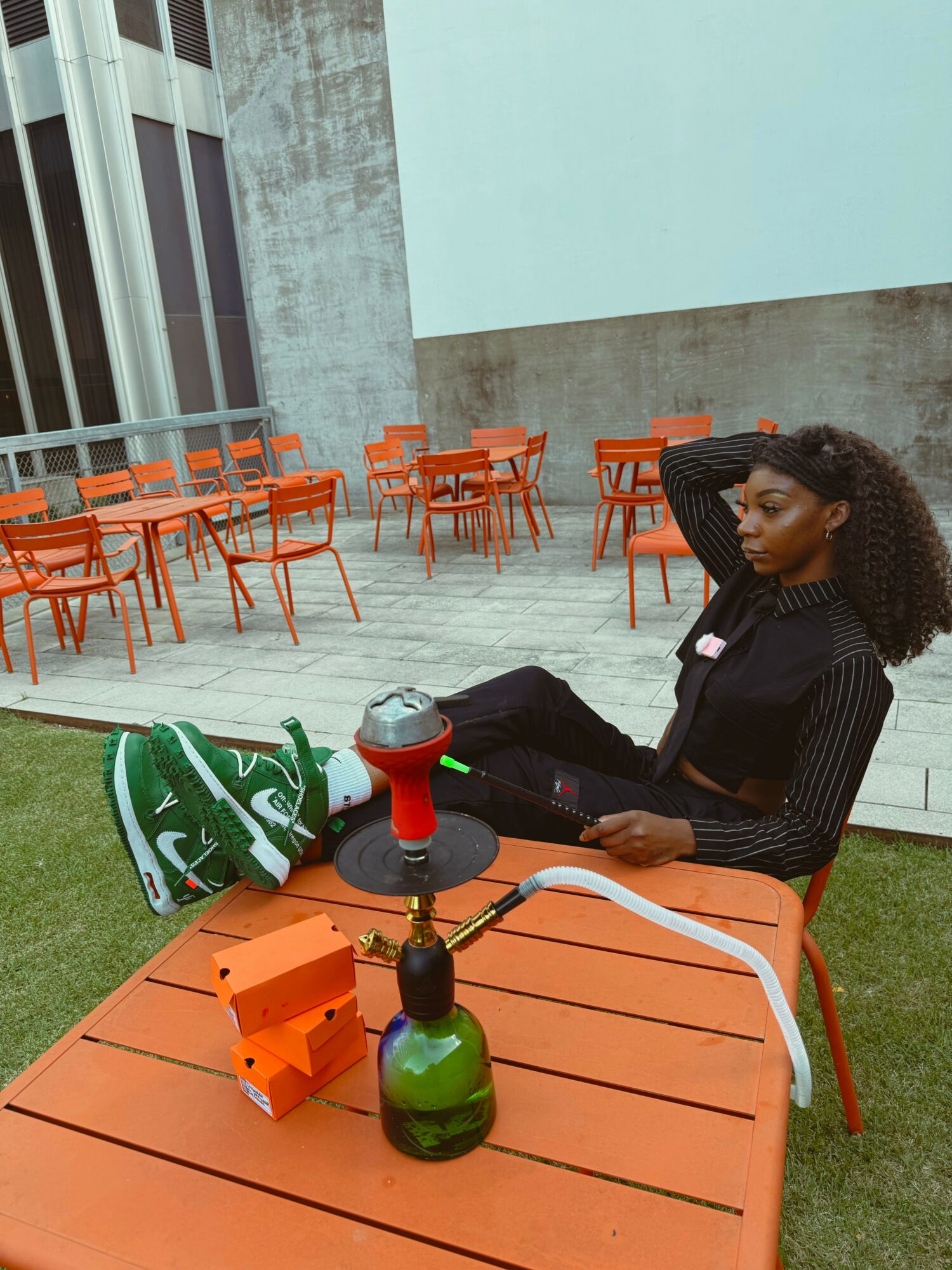 Young woman with curly hair sitting at an orange table outdoors, using a laptop, with a building and orange chairs in background.