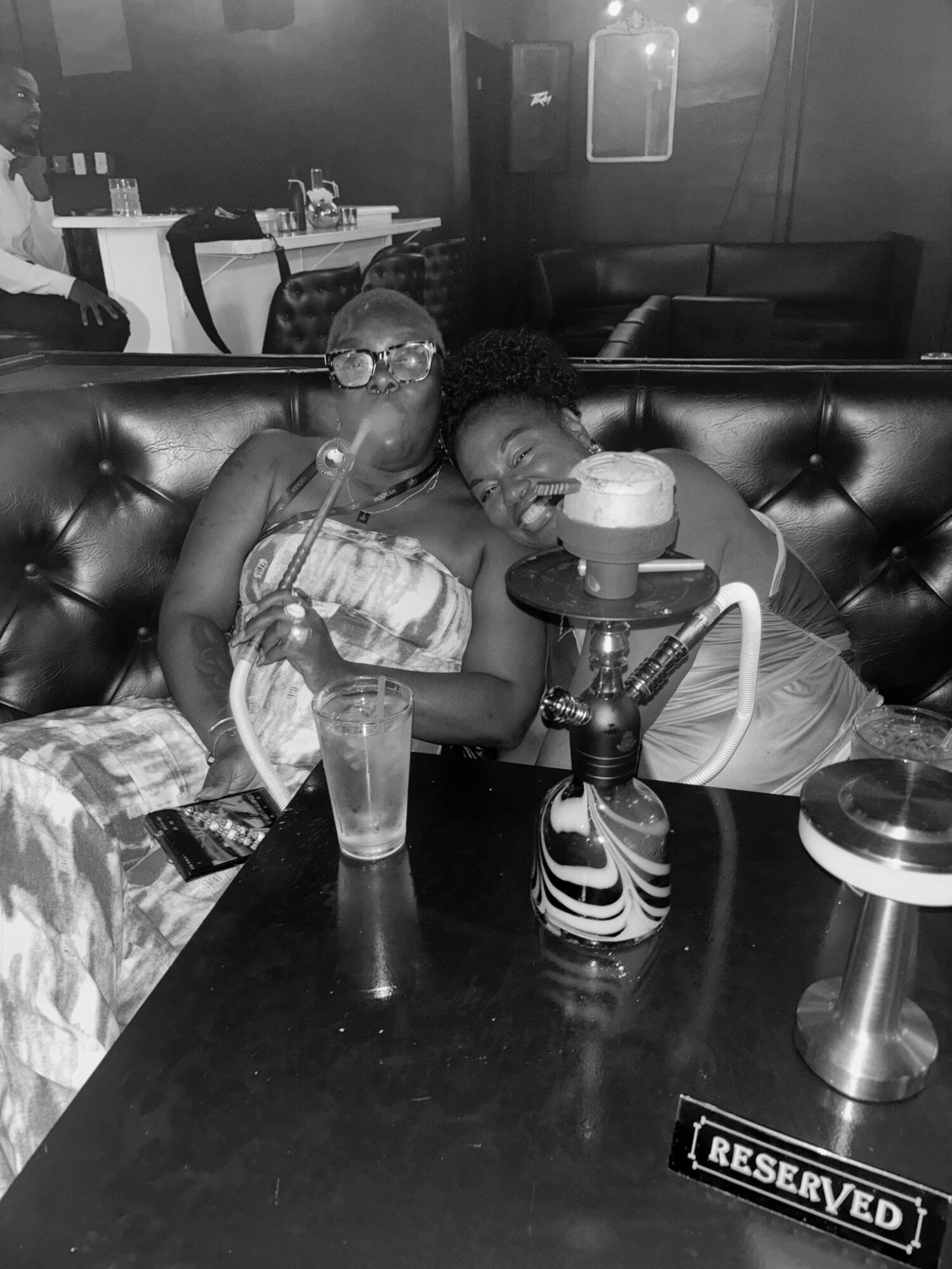 Two women sit close together on a booth in a dimly lit restaurant, sharing a hookah and drinks on the table.