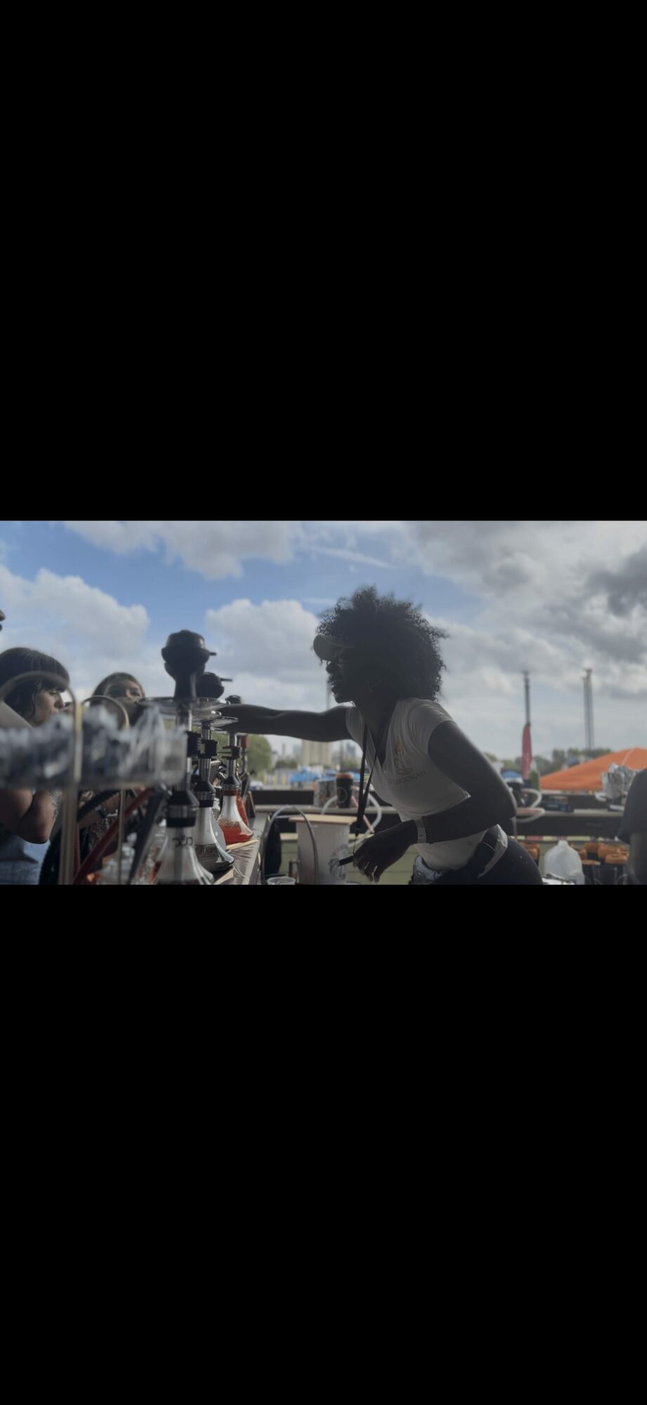 Two people with curly hair interact outdoors with a camera and equipment, under a partly cloudy sky.