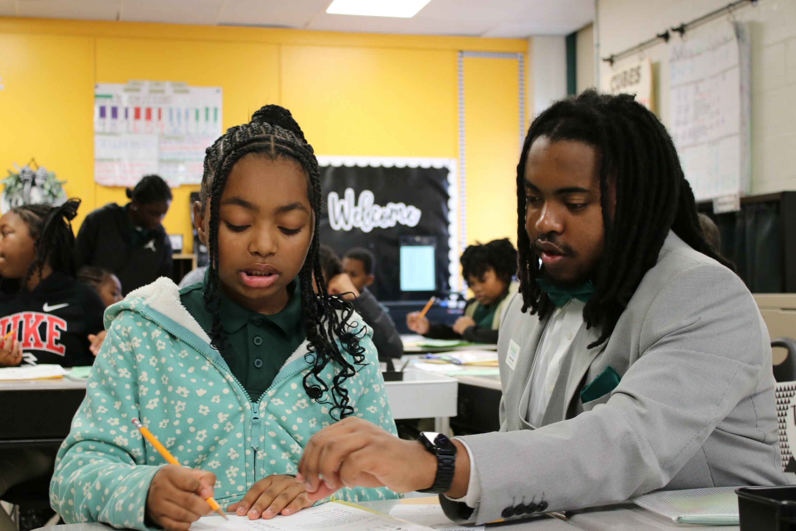 A young girl and a man sit at a desk in a classroom, working together on a task, with other students in the background.