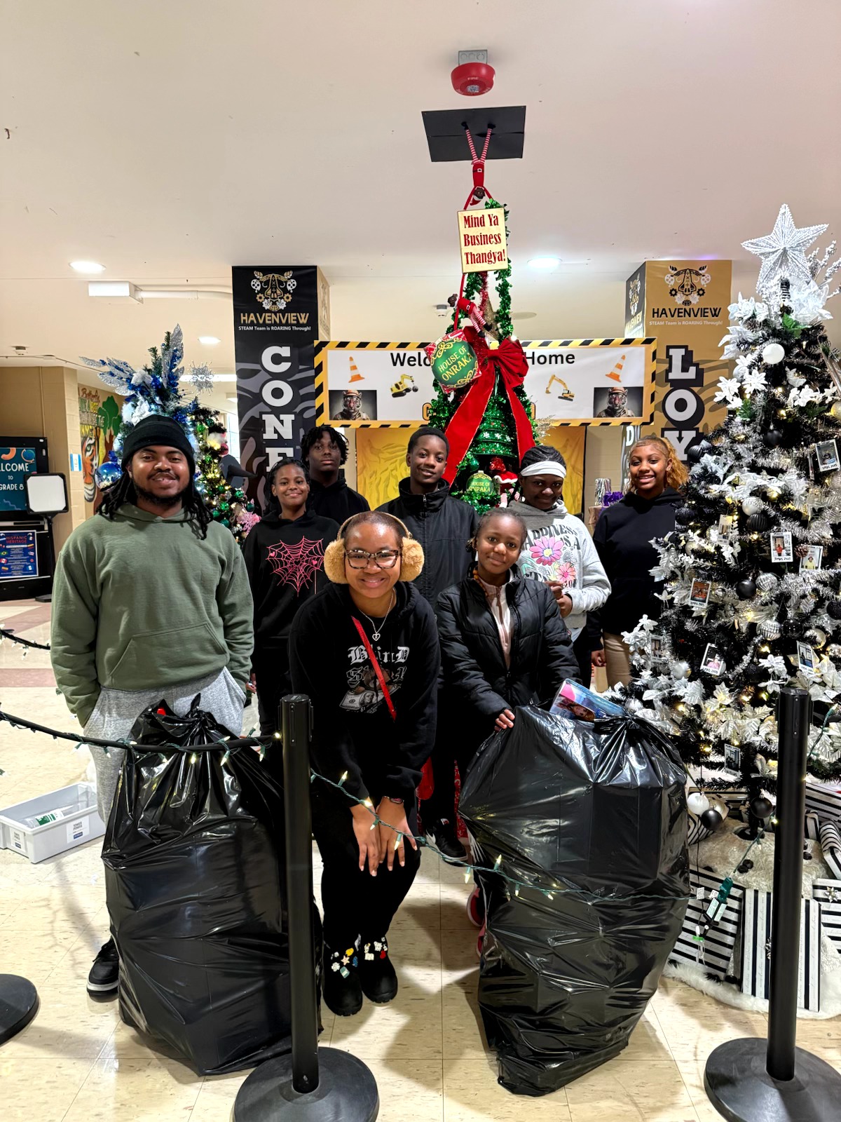 Group of six people standing in a store with Christmas decorations, holding large black trash bags, smiling, with a decorated Christmas tree behind them.