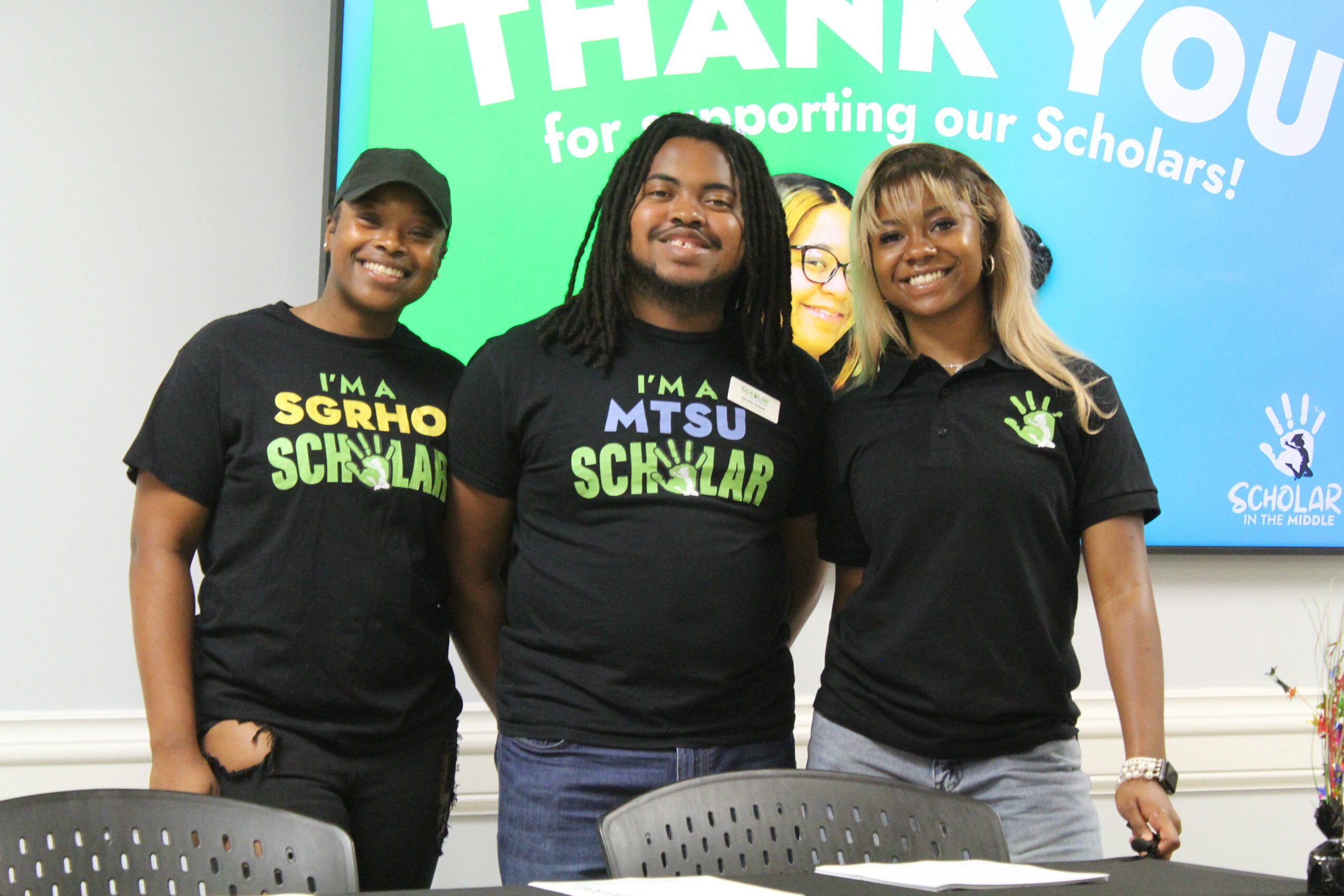 Three people standing in front of a screen, smiling, wearing black shirts with green and white text, in a room.