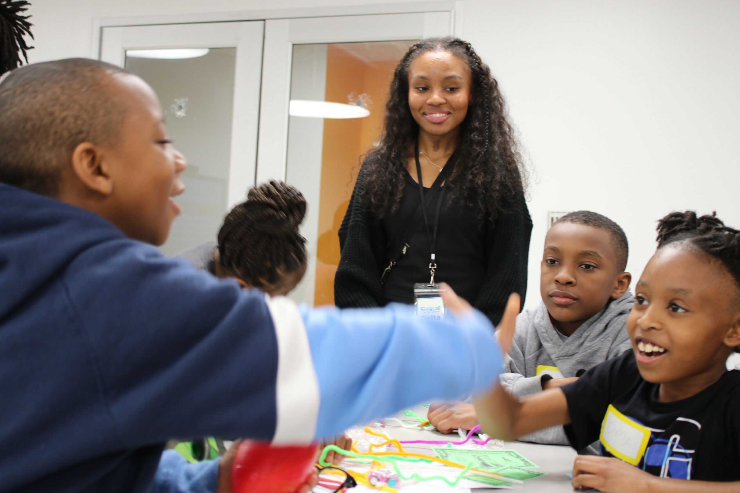 Group of children and a woman smiling and interacting in a classroom or playroom, with colorful toys on a table.