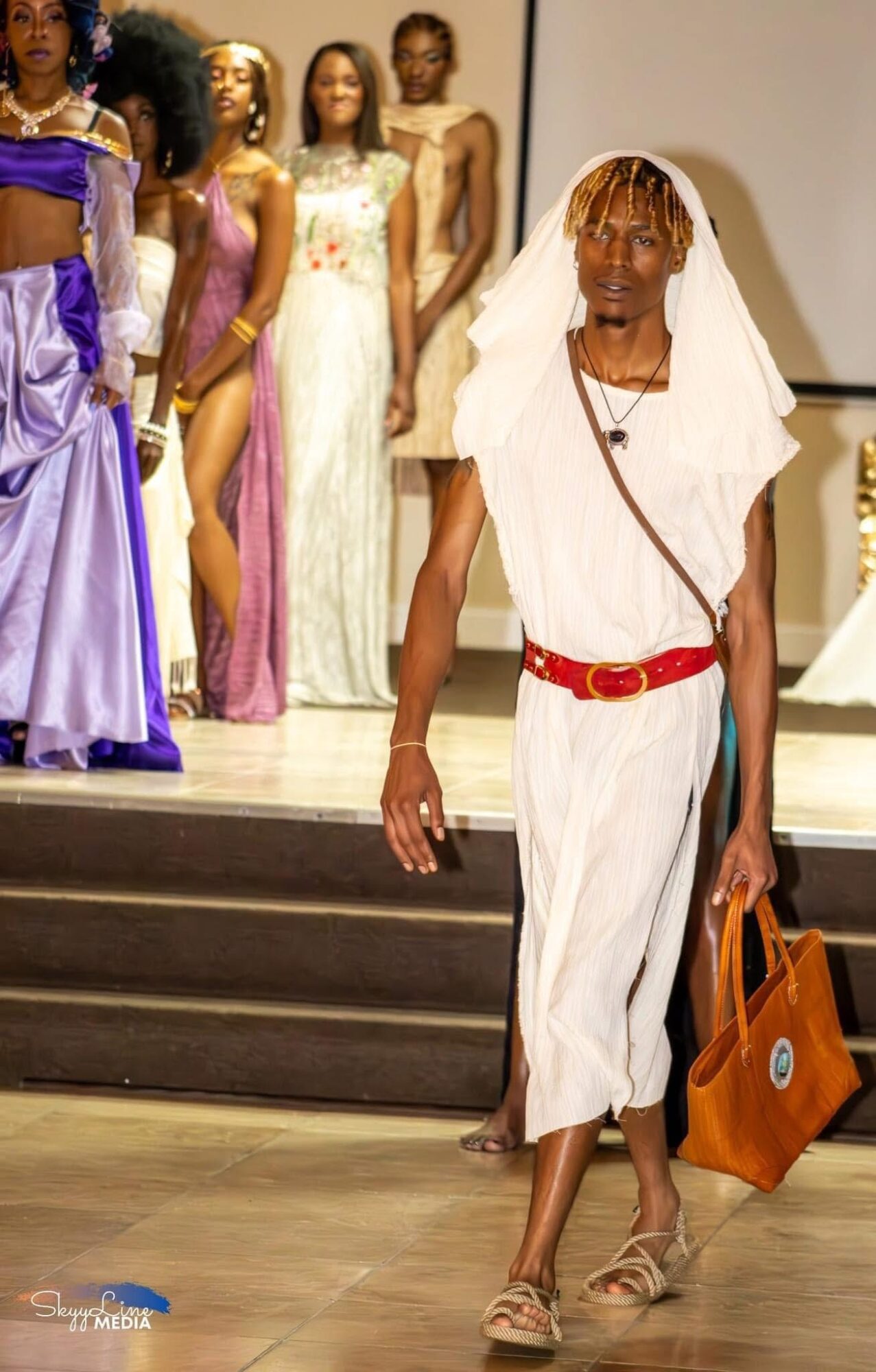 Woman walking in front of a runway with models in colorful dresses, carrying a large bag, wearing a white outfit and head covering.