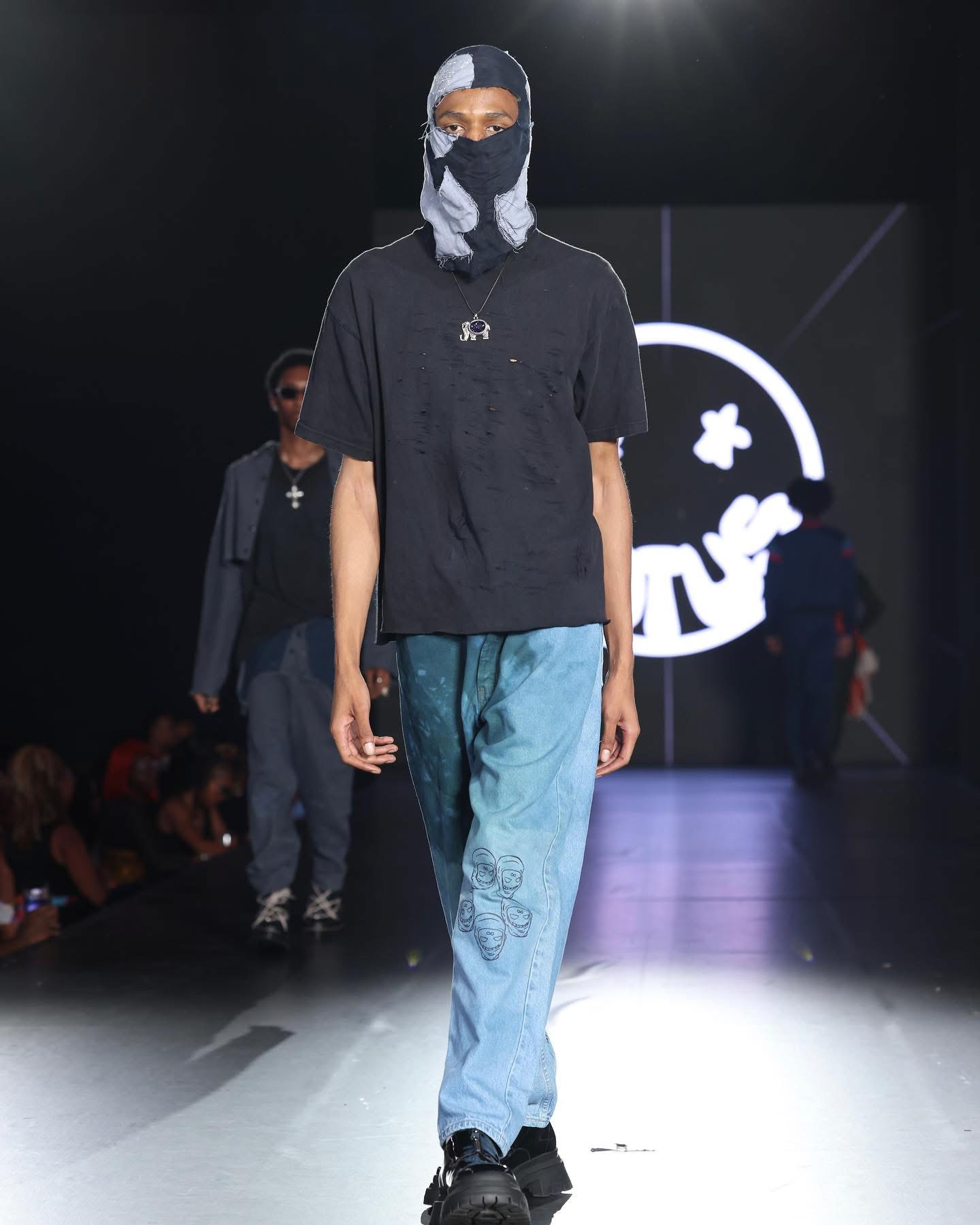 Person walking on runway wearing black shirt, blue pants, face mask, and head covering, with a logo in the background.