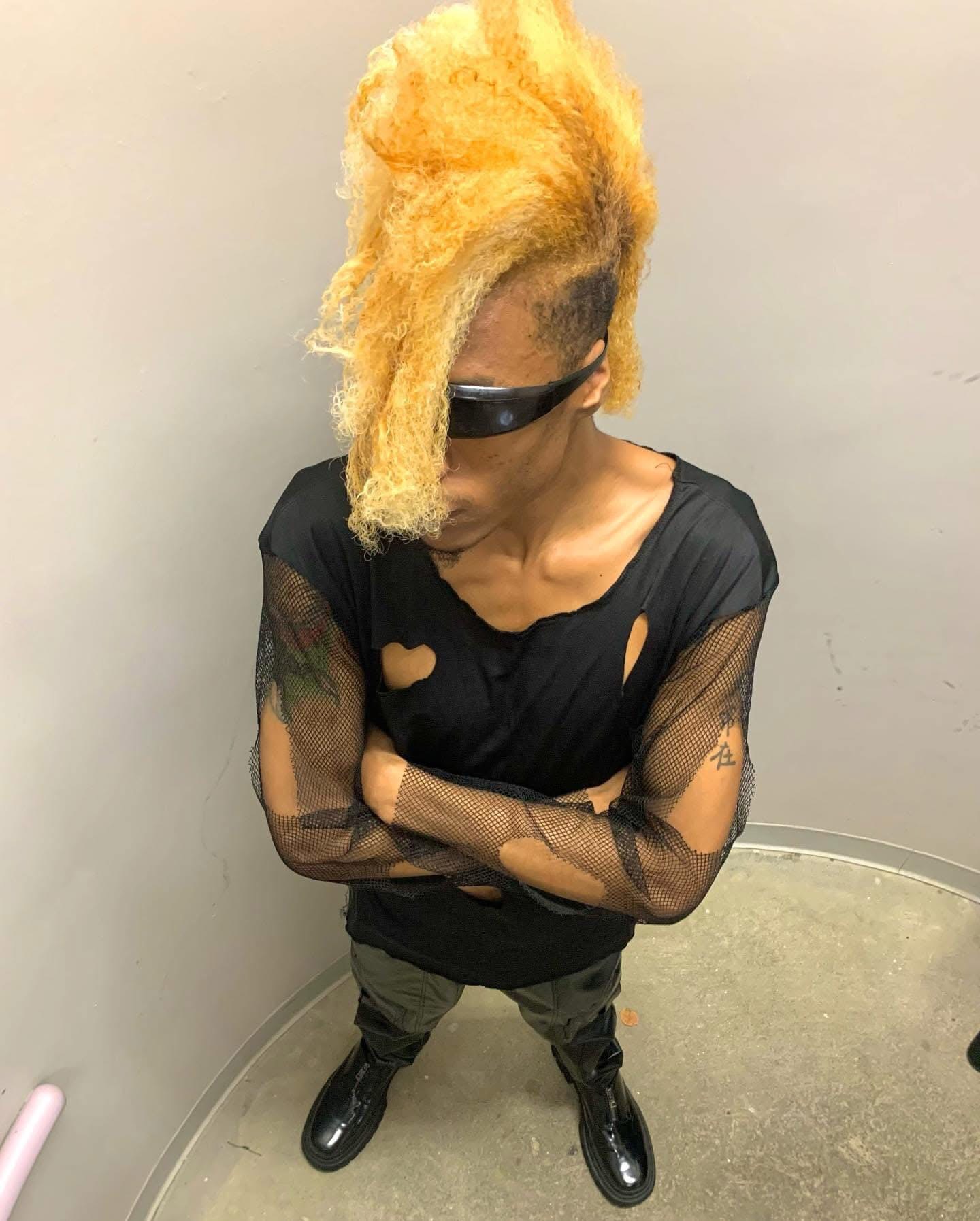 Person with bright yellow hair, wearing sunglasses, black torn shirt, and camouflage pants, standing with arms crossed against a plain wall.