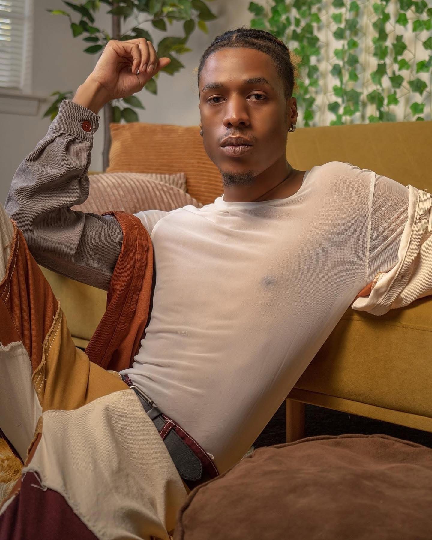 Person sitting on a couch with a serious expression, wearing a light-colored shirt and a jacket, in a room with green plants.