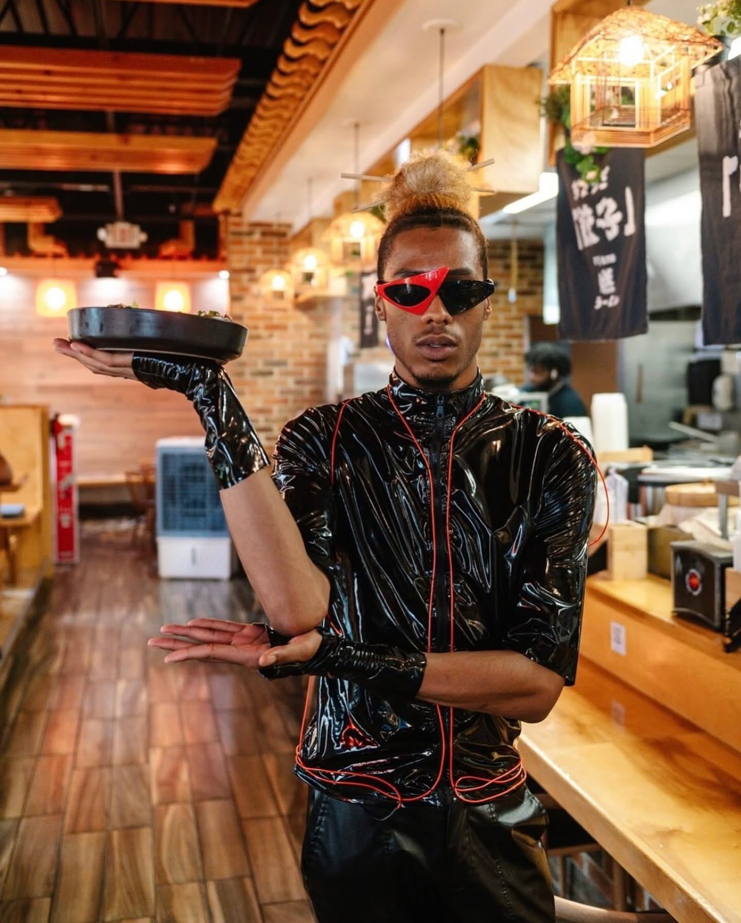 Person with styled hair, wearing red sunglasses, black shiny outfit, and gloves, standing in a restaurant or cafe interior.