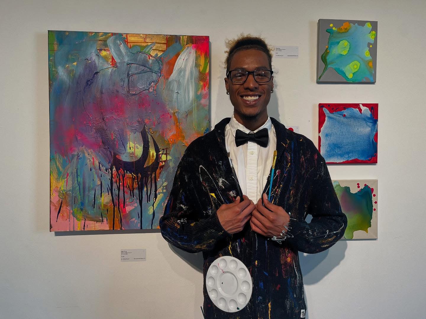 Young man in formal attire with a bow tie, smiling, standing in front of colorful abstract paintings.