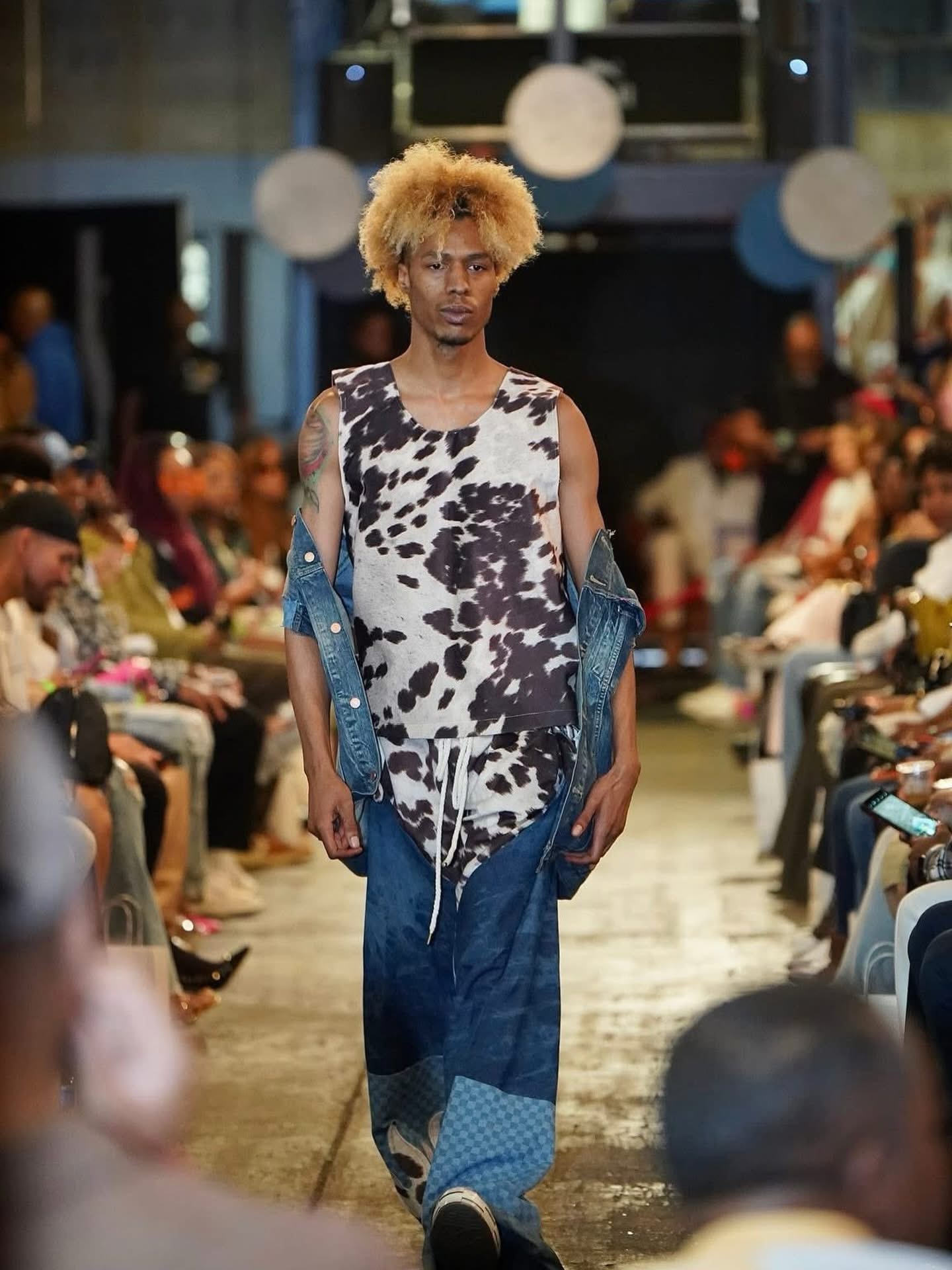 Female model walking down runway in patterned dress and blue outerwear, audience seated on both sides, indoor setting.