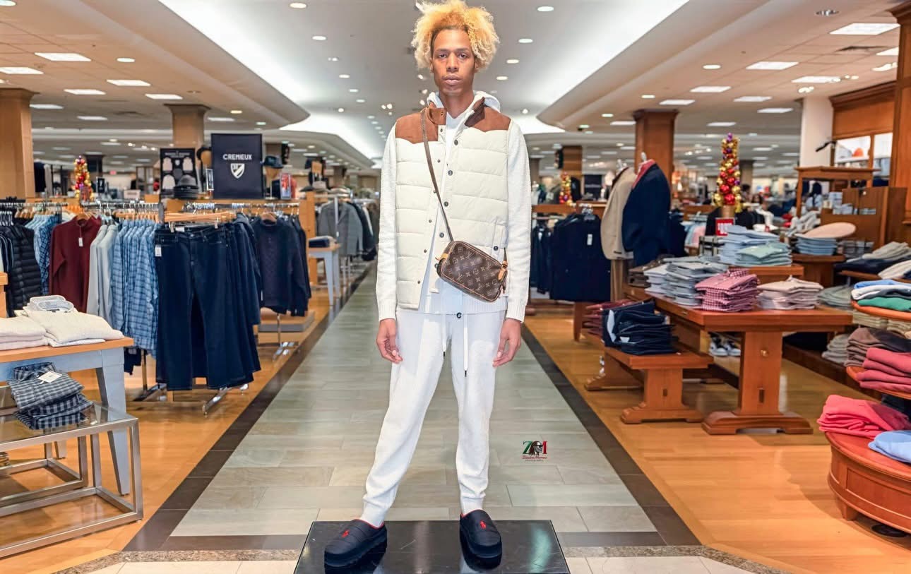 Person with blonde hair wearing white outfit and brown crossbody bag standing in clothing store aisle.