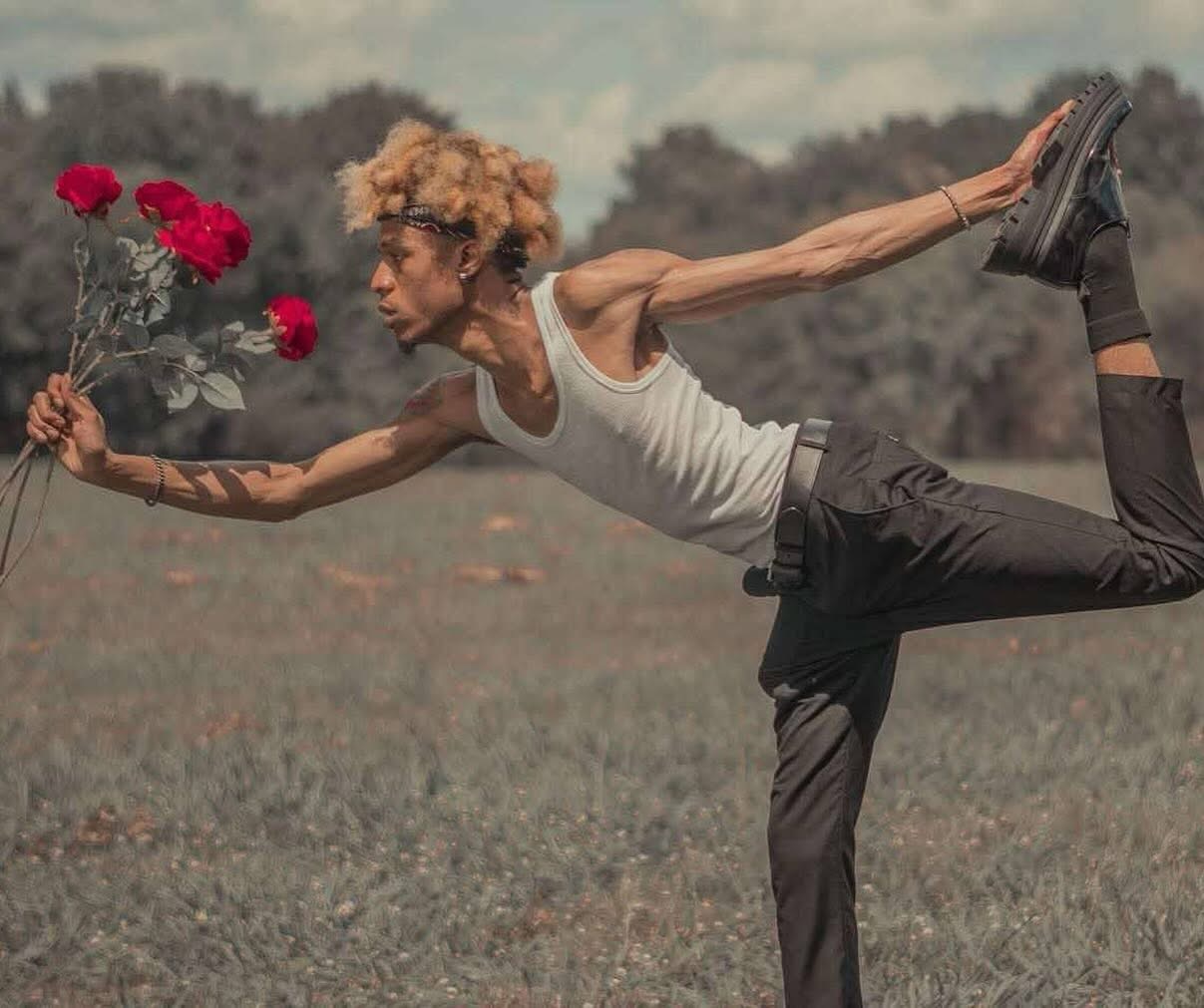 Person balancing on one leg holding flowers, outdoors, with arms extended and head turned sideways.