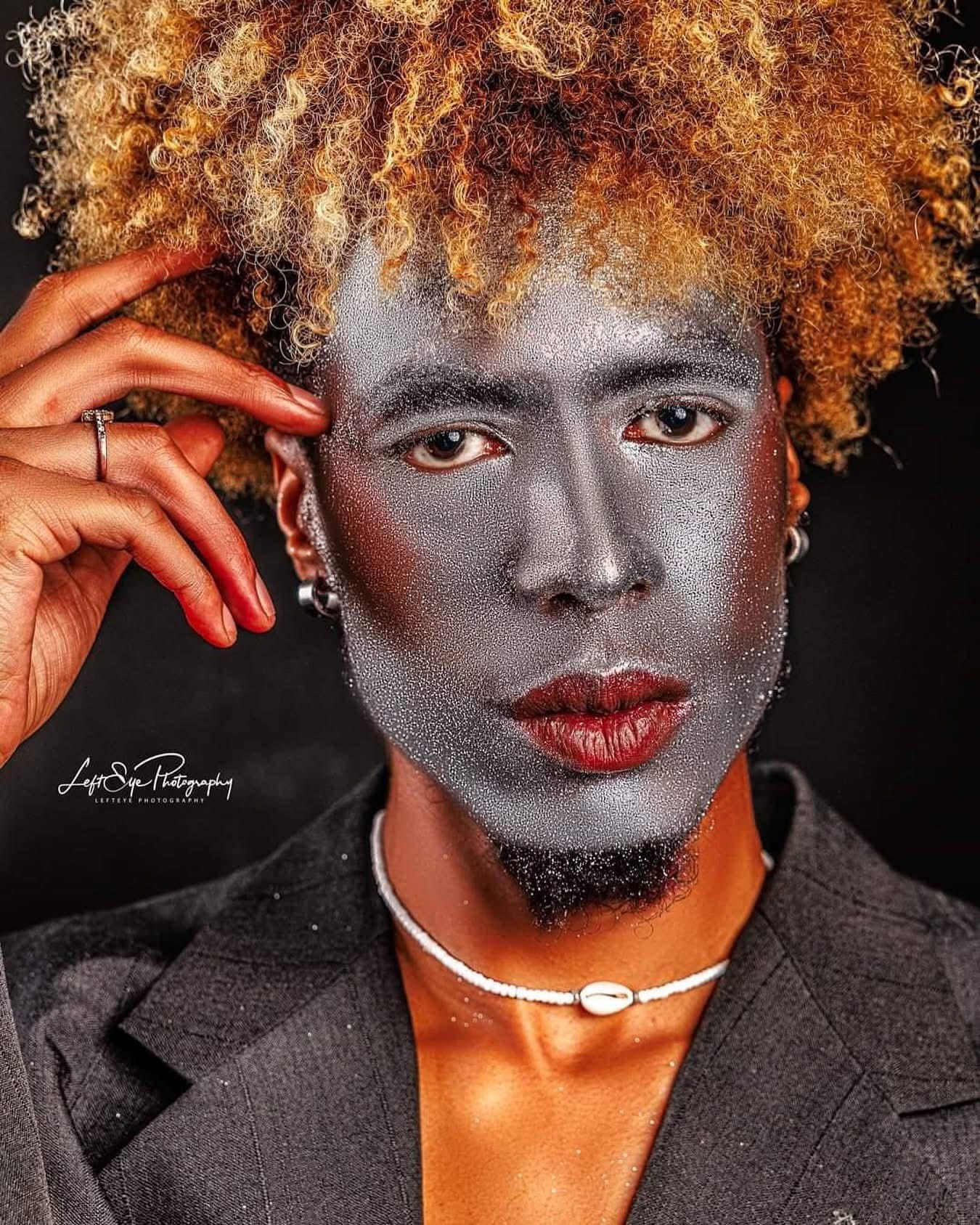 Person with curly blonde hair, silver face makeup, red lipstick, wearing earrings, necklace, and dark blazer.